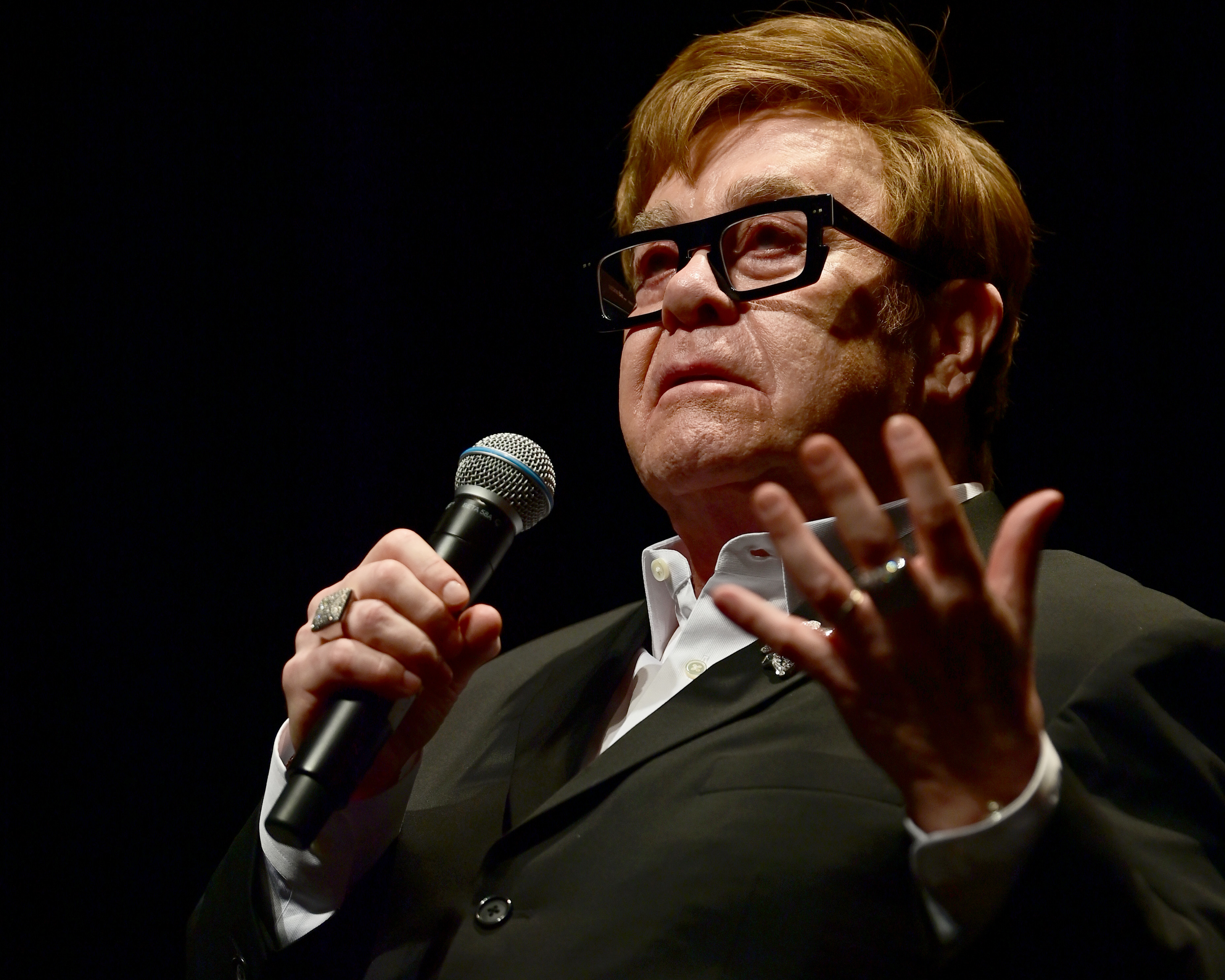Elton John is holding a microphone and speaking on stage. He is wearing a black suit with a white shirt and black rectangular glasses