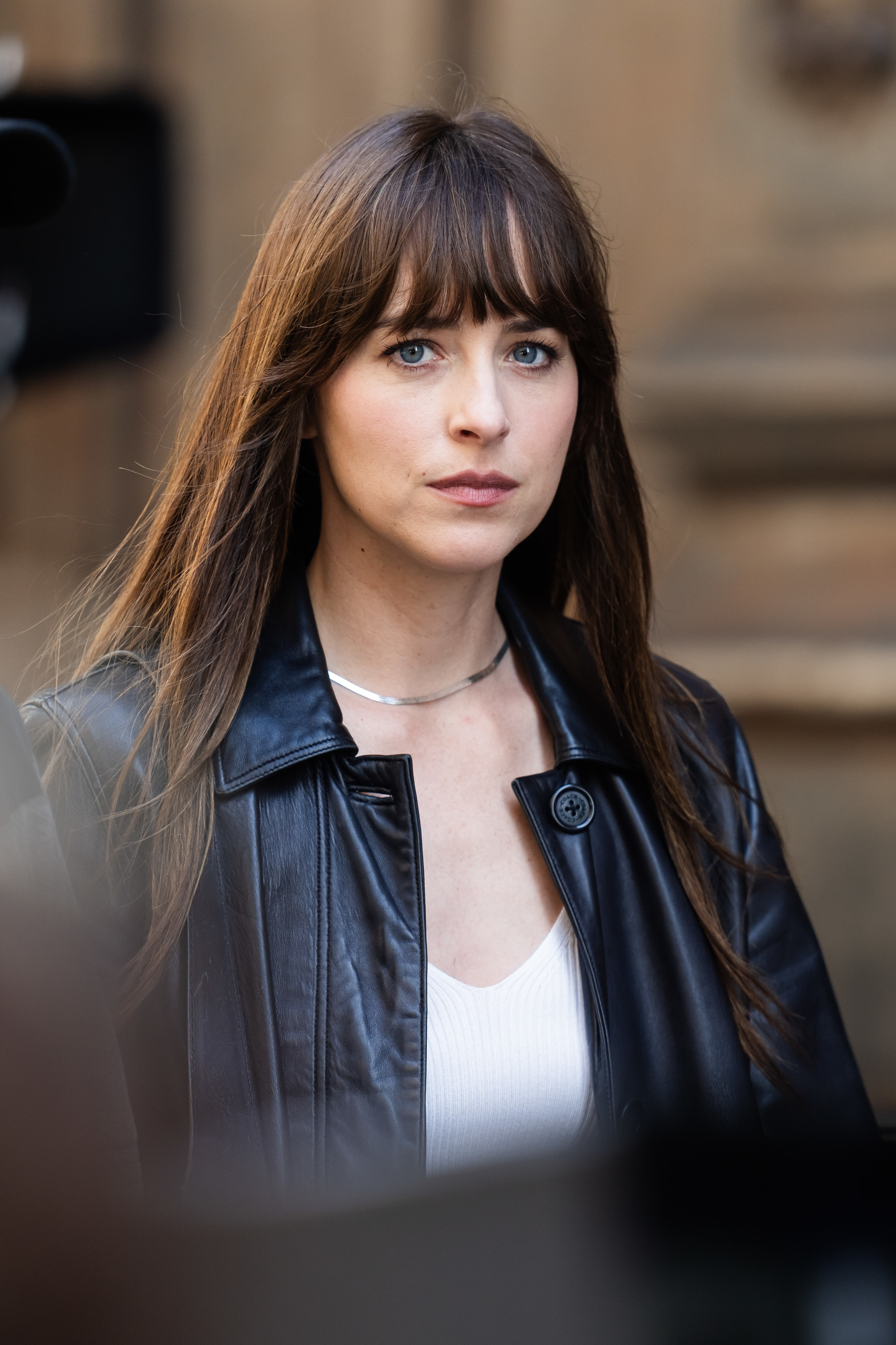 Dakota Johnson wearing a black leather jacket over a white top, looking at the camera