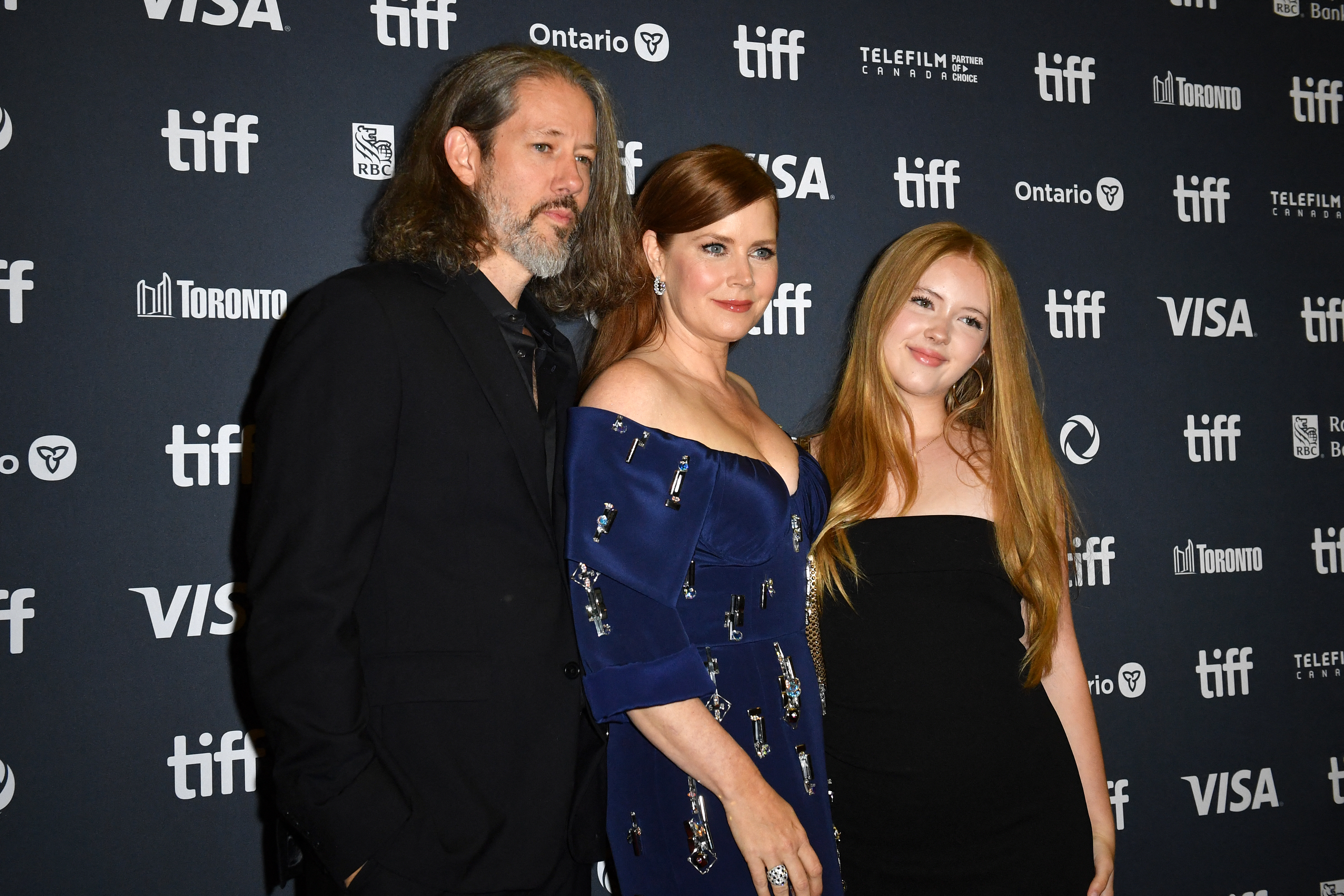 Three people pose on the TIFF red carpet: a man in a black suit, Amy Adams in an elegant blue dress, and a young woman in a black dress