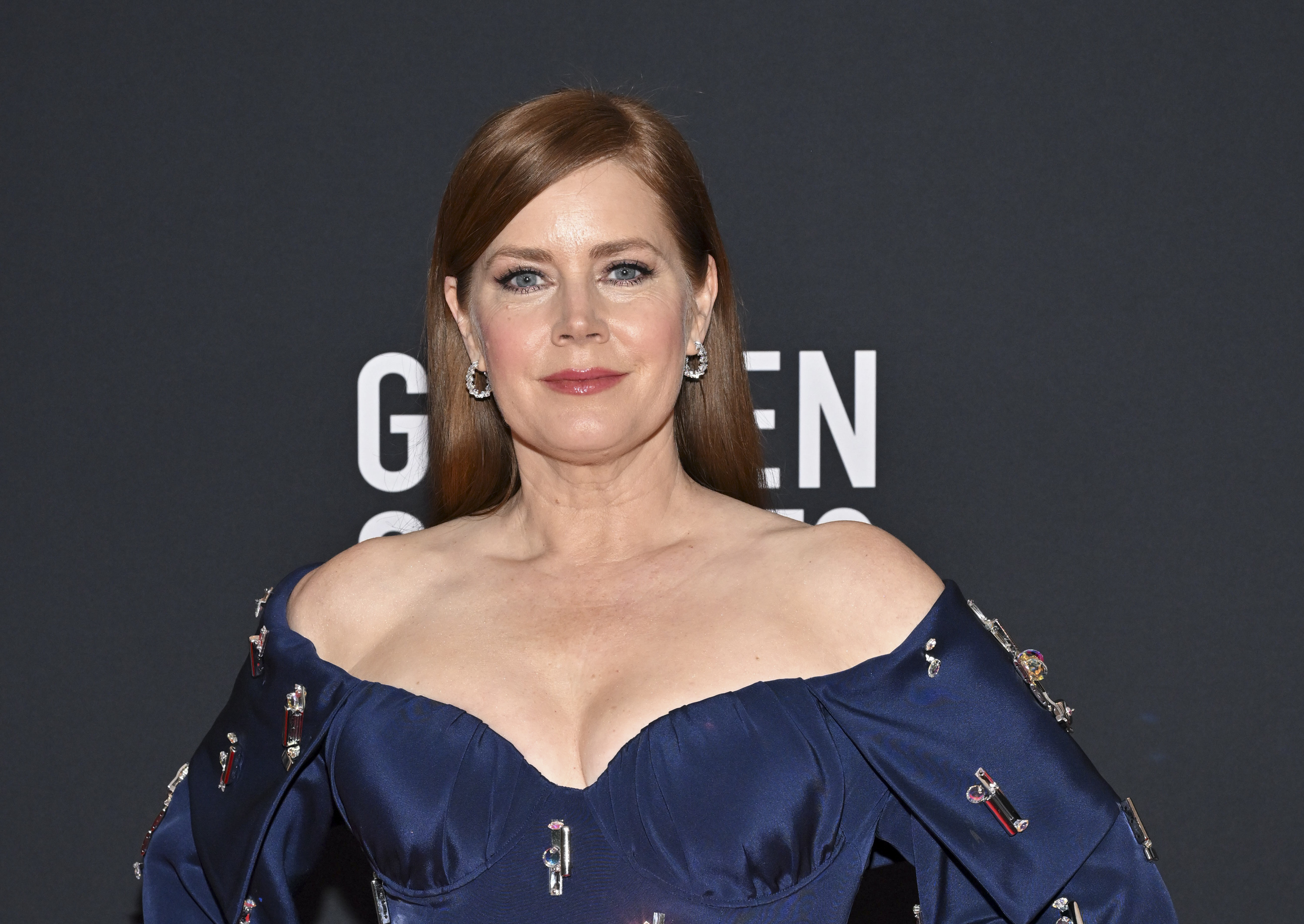 Amy Adams in an off-the-shoulder, embellished navy gown at a formal event, posing in front of a backdrop with text partially visible