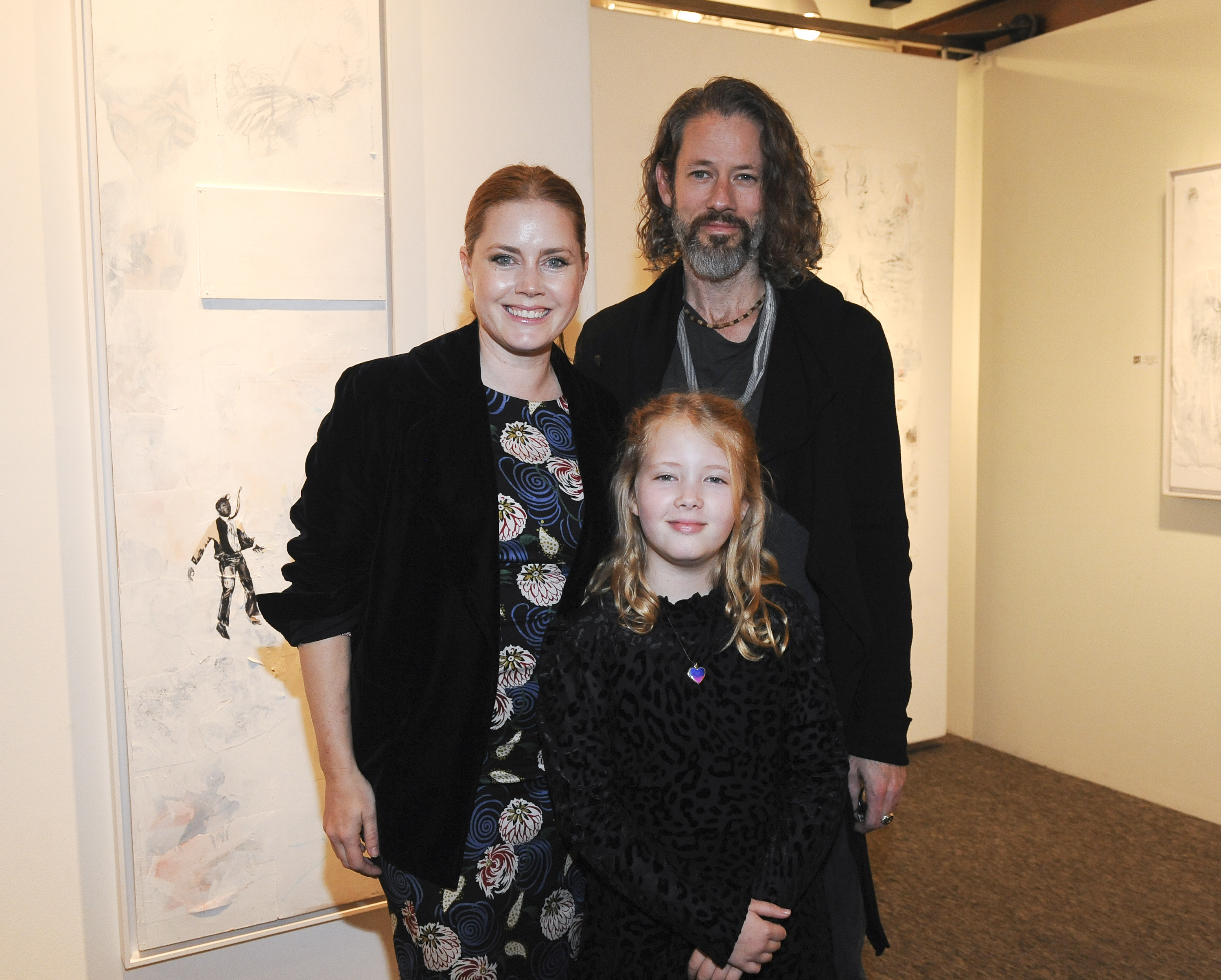 Amy Adams and Darren Le Gallo stand with their daughter in an art gallery. Amy wears a patterned dress under a dark jacket, Darren wears a dark outfit