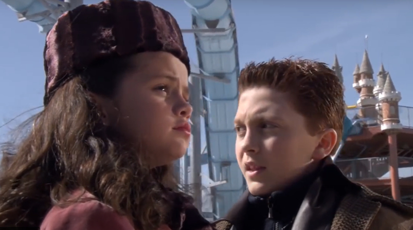 Selena Gomez and Daryl Sabara in winter clothing, standing outside a theme park with castle structures in the background, appearing focused