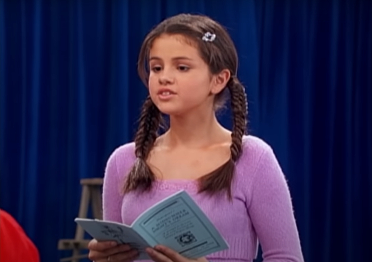 Selena Gomez, wearing a long-sleeved top with two braids, holds a blue booklet and speaks in front of a blue curtain backdrop