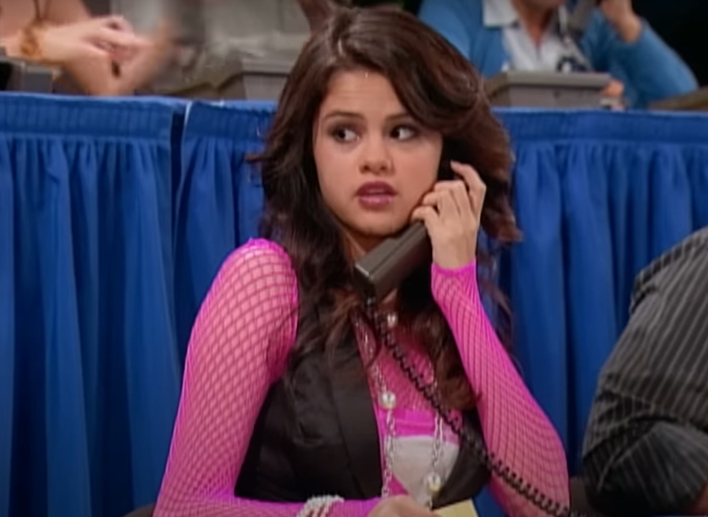 Selena Gomez in a scene from &quot;Hannah Montana,&quot; sitting at a table and holding a phone. She is wearing a pink mesh top under a black vest