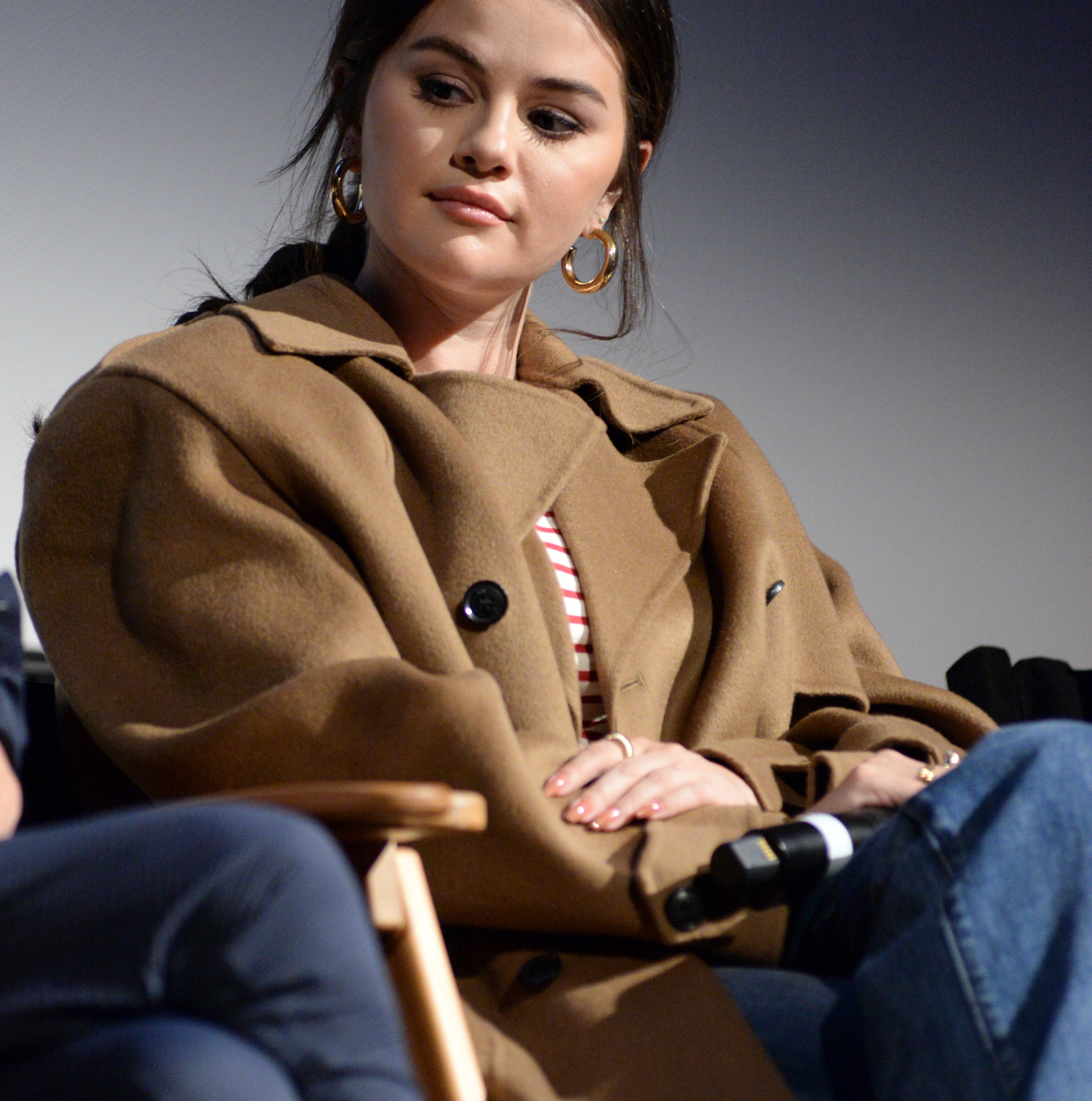 Selena Gomez in a casual outfit with a brown coat and jeans, sitting on a chair during an event, holding a microphone