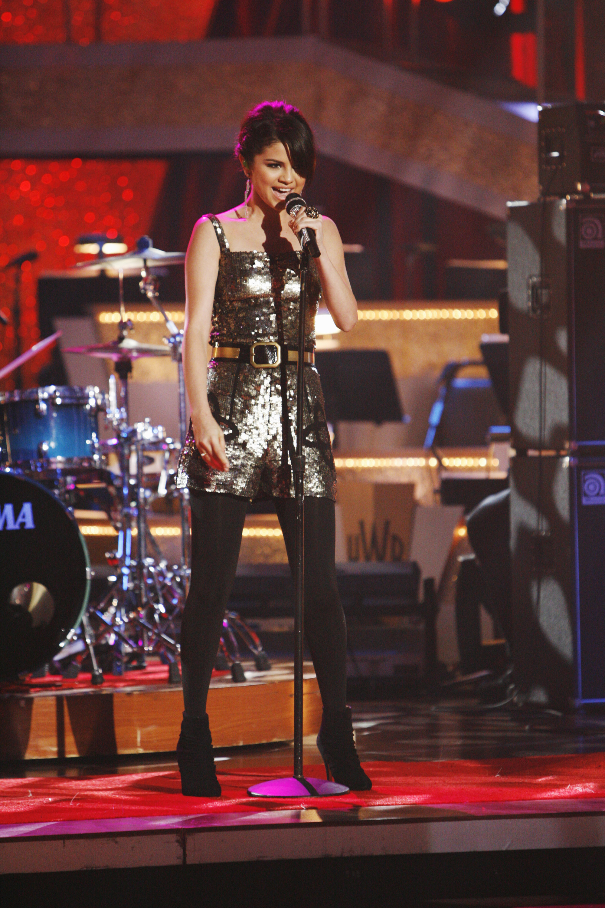 Selena Gomez on stage, wearing a sequined dress and black tights, singing into a microphone with a band setup in the background