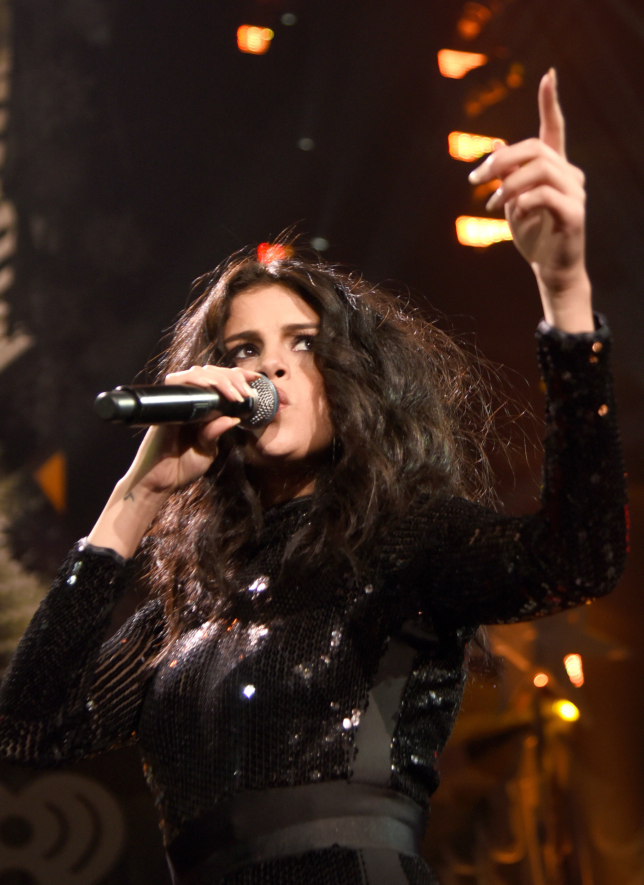 Selena Gomez performs on stage, holds a microphone, and points upward. She wears a sparkly, long-sleeved dress