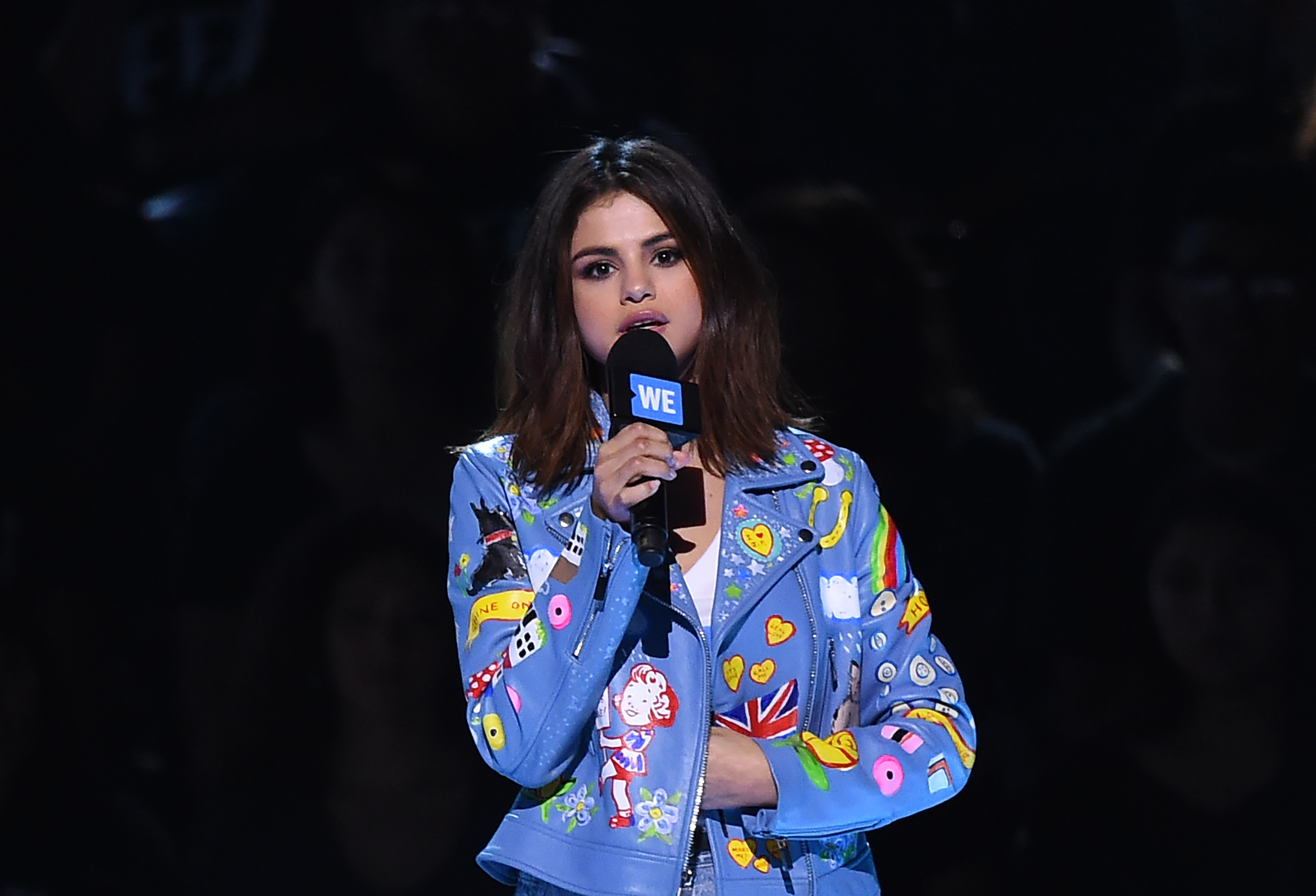 Selena Gomez speaks on a dark stage at a WE Day event, holding a WE-branded microphone. She wears a colorful, patchwork jacket with various fun designs