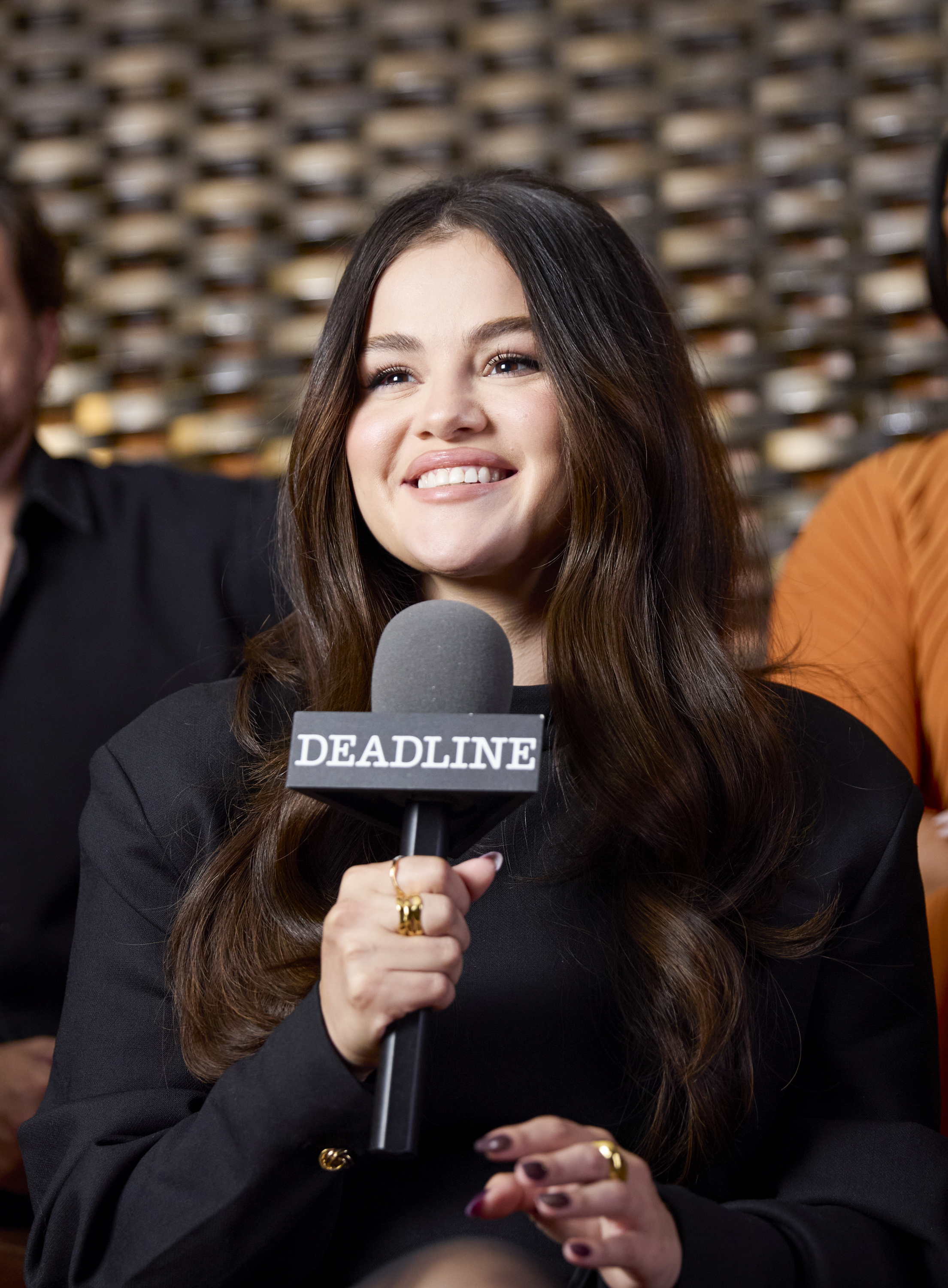 Selena Gomez smiles while holding a &quot;DEADLINE&quot; microphone during an interview, wearing a black outfit with long, wavy hair