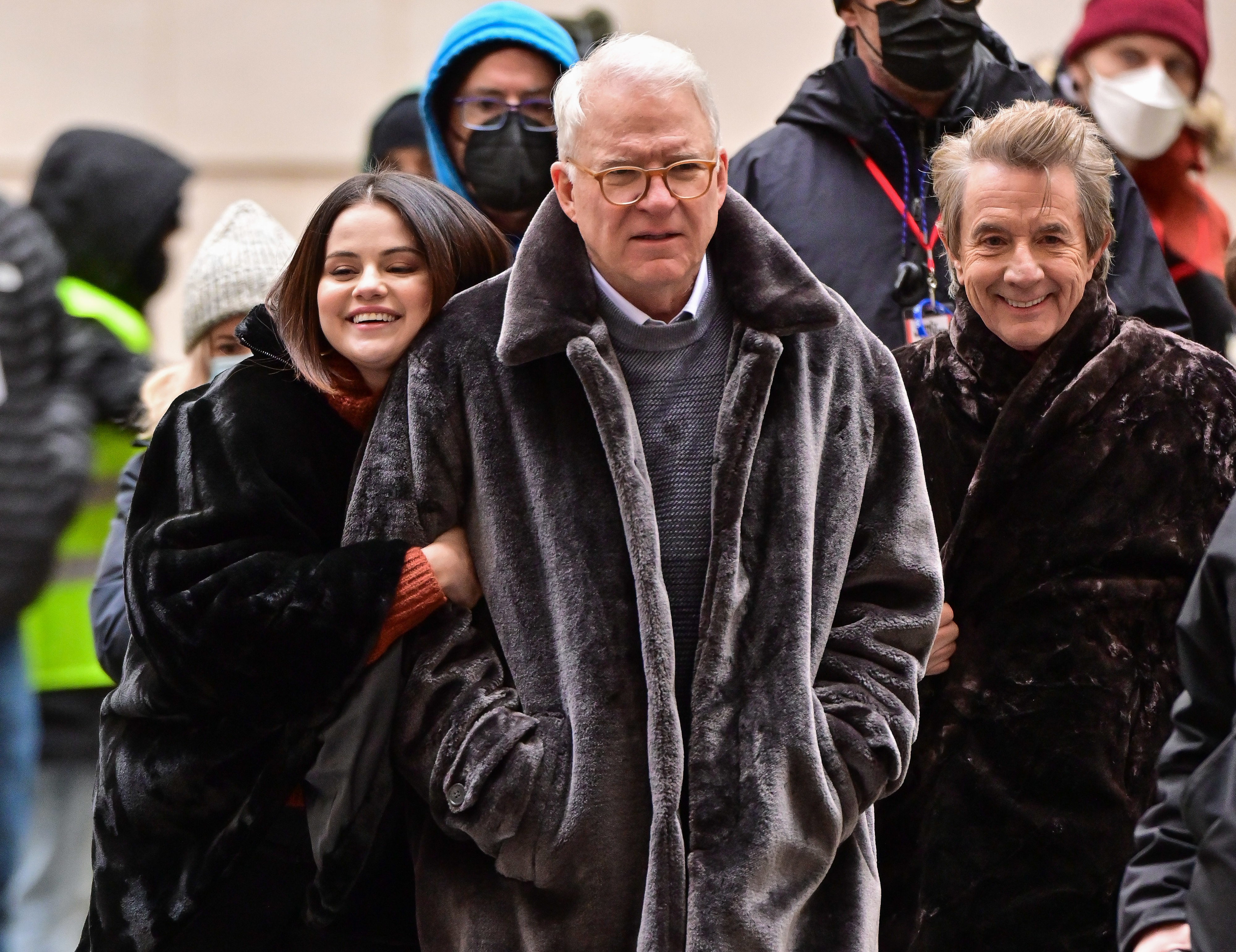 Selena Gomez, Steve Martin, and Martin Short smiling and wearing winter coats while walking outdoors among people, including some in masks