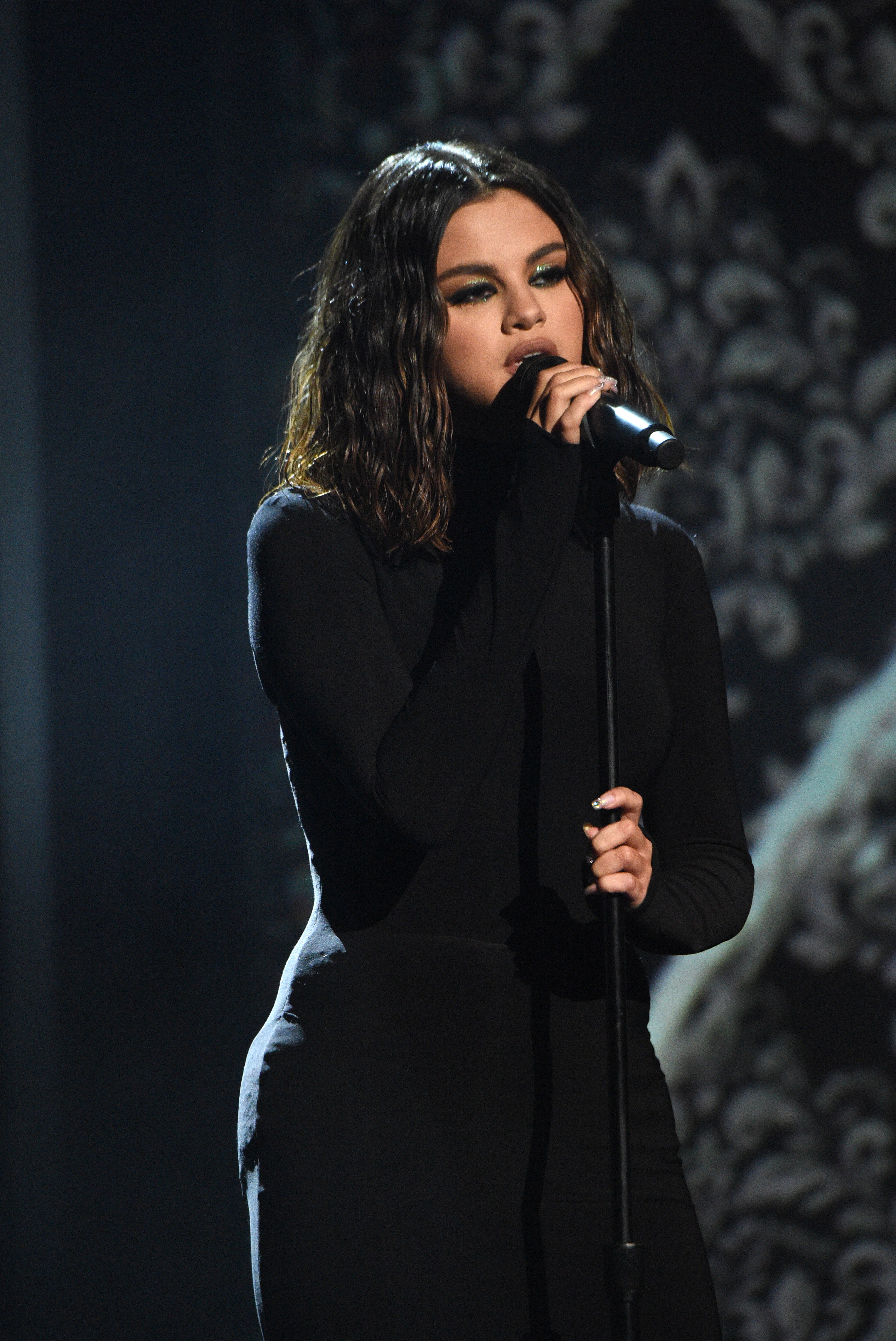 Selena Gomez is on stage, wearing a fitted black dress, holding a microphone and performing against an ornate background