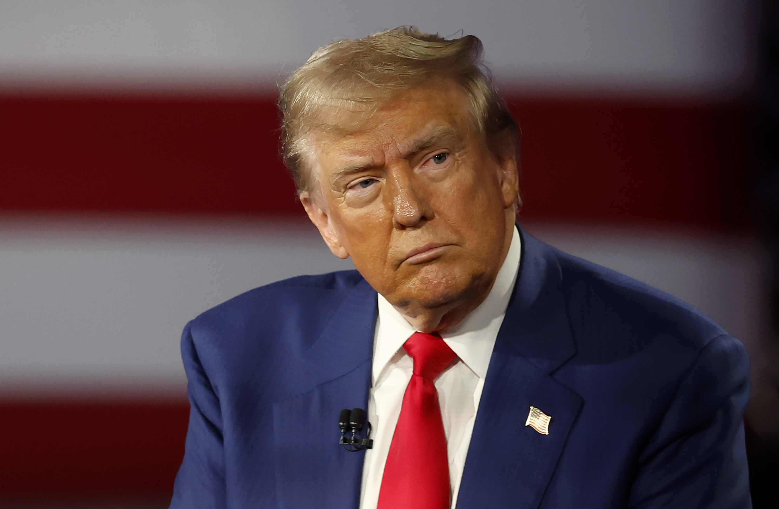 Donald Trump, wearing a suit and tie, attentively listens during an event with an American flag in the background