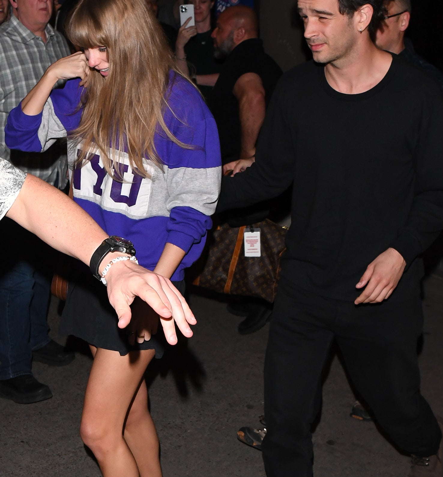 Taylor Swift, wearing a casual sweatshirt and skirt, walks with Matty Healy, dressed in a black outfit, through a crowd of fans and photographers