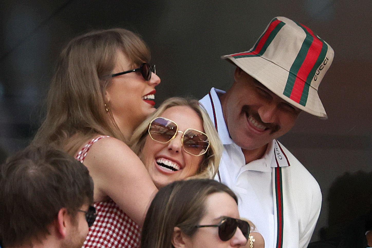 Taylor Swift, Brittany Mahomes and Travis Kelce share a joyful moment while embracing. Travis wears a stylish bucket hat and blazer