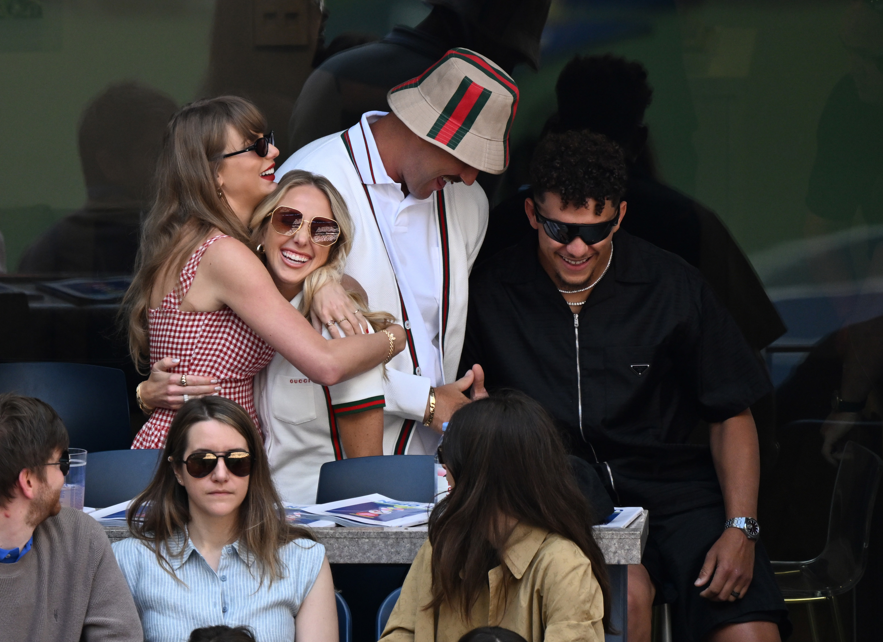 Taylor Swift, Brittany Mahomes and Travis Kelce share a joyful moment while embracing. Travis wears a stylish bucket hat and blazer