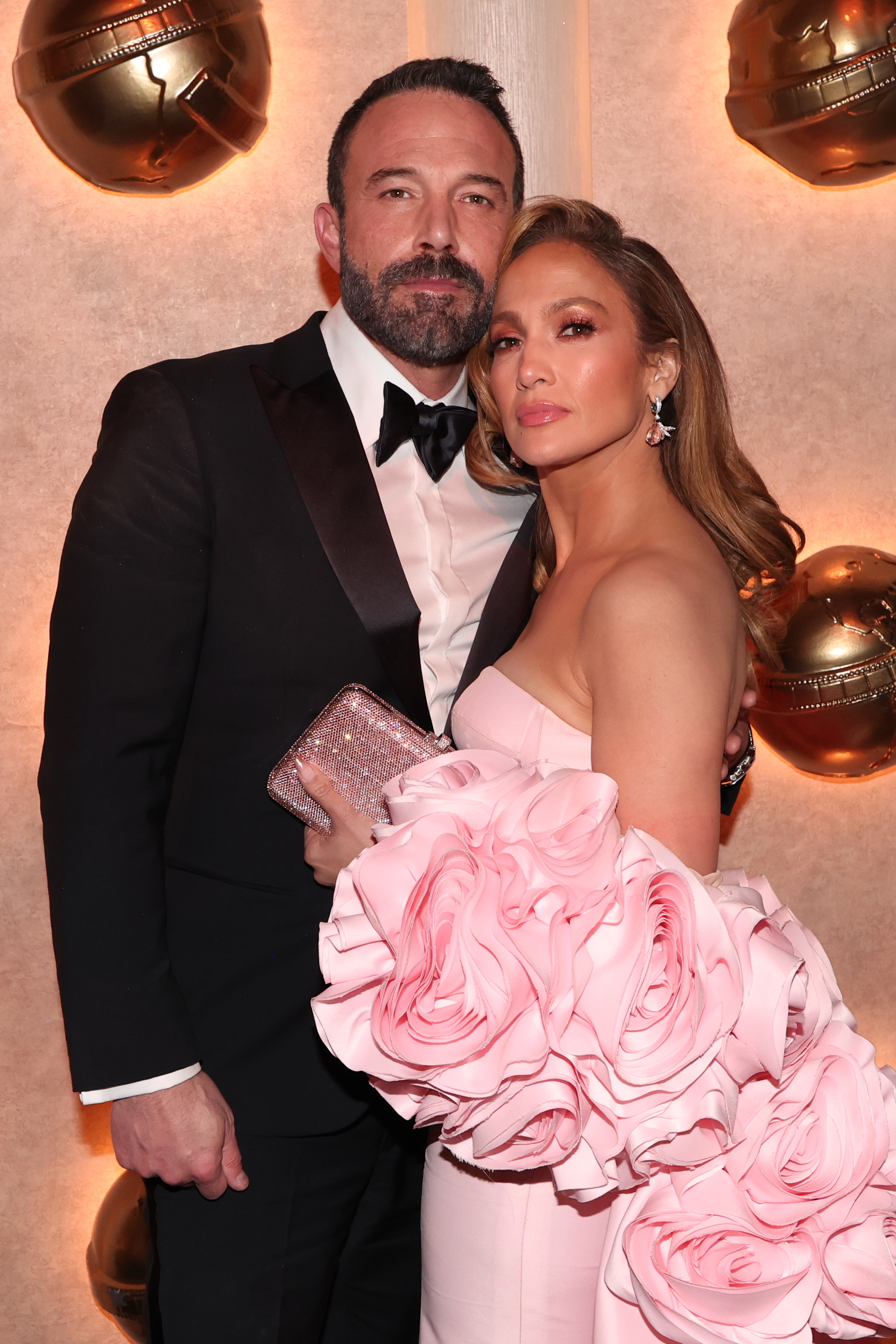 Ben Affleck and Jennifer Lopez pose for a photo. Ben wears a classic tuxedo and Jennifer wears a strapless gown with large floral accents