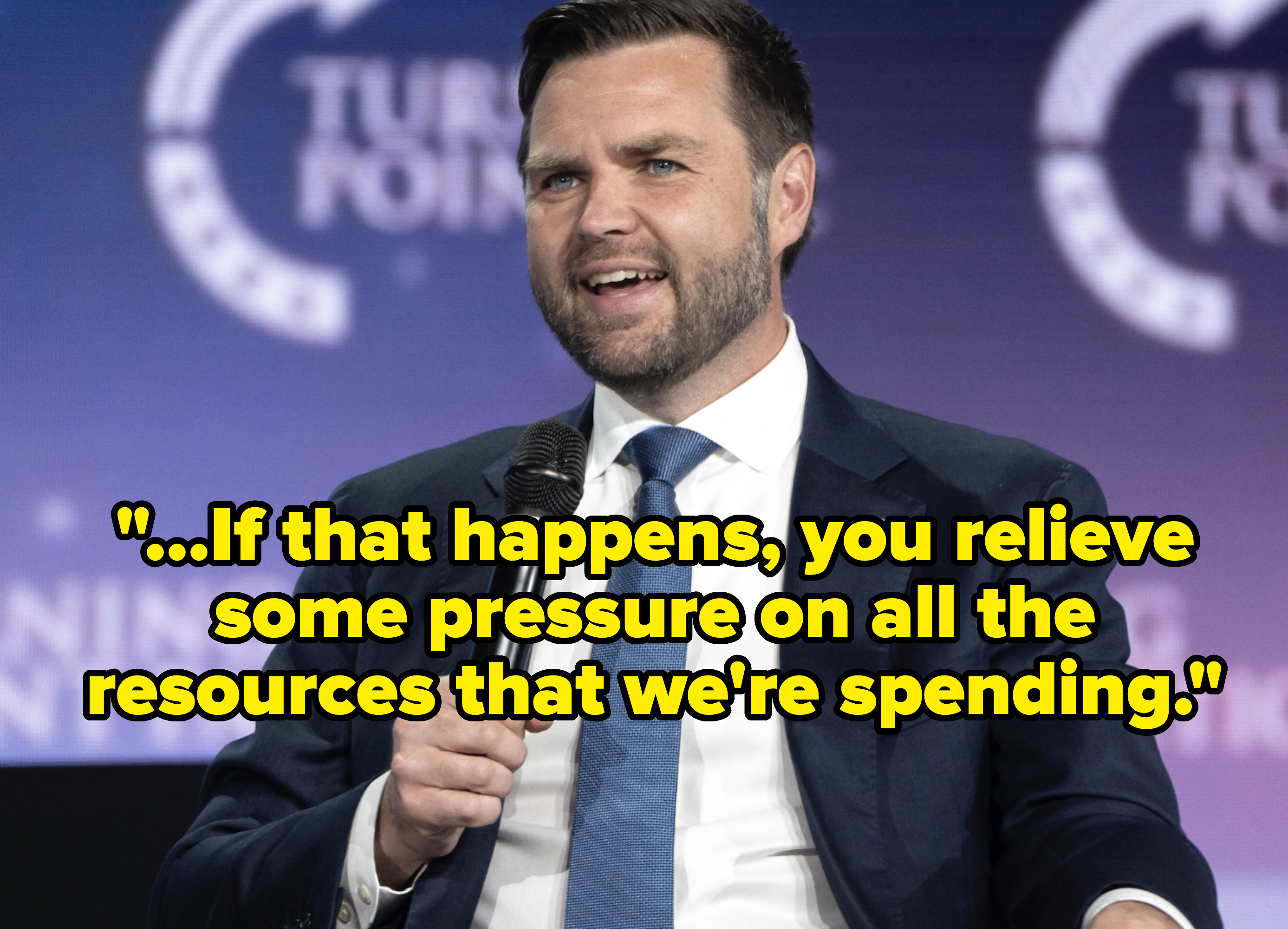 J.D. Vance speaks animatedly into a microphone at a Turning Point USA event, wearing a suit and tie with a small U.S. flag pin on the lapel