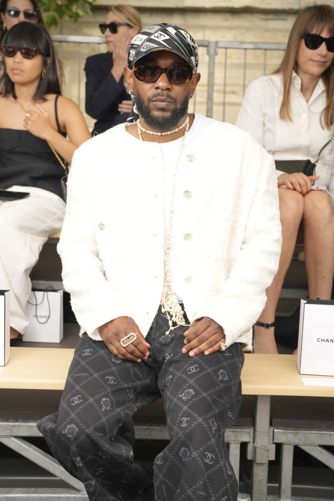 Kendrick Lamar sitting front row at a fashion show, wearing a jacket, patterned pants, and a headscarf. People in the background wear sunglasses