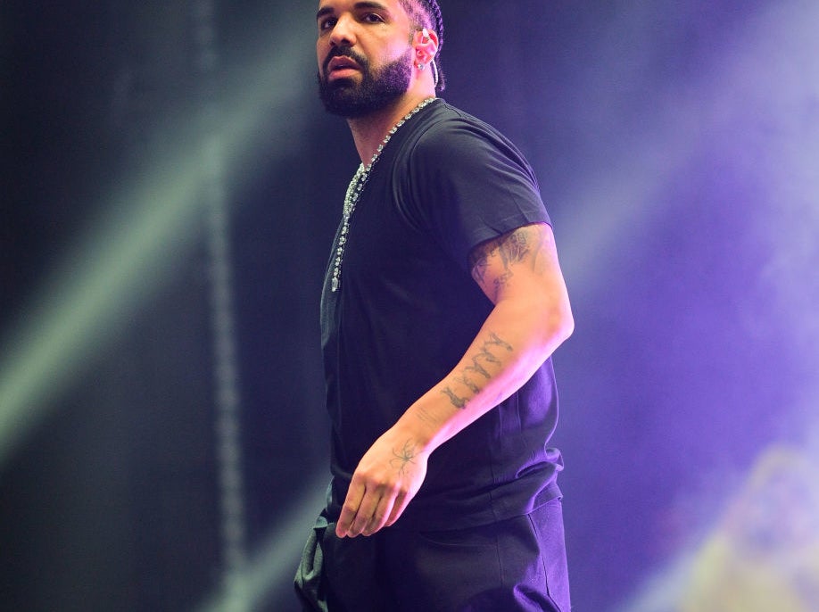 Drake performs on stage wearing a black outfit with a necklace, looking to the side amidst bright stage lights