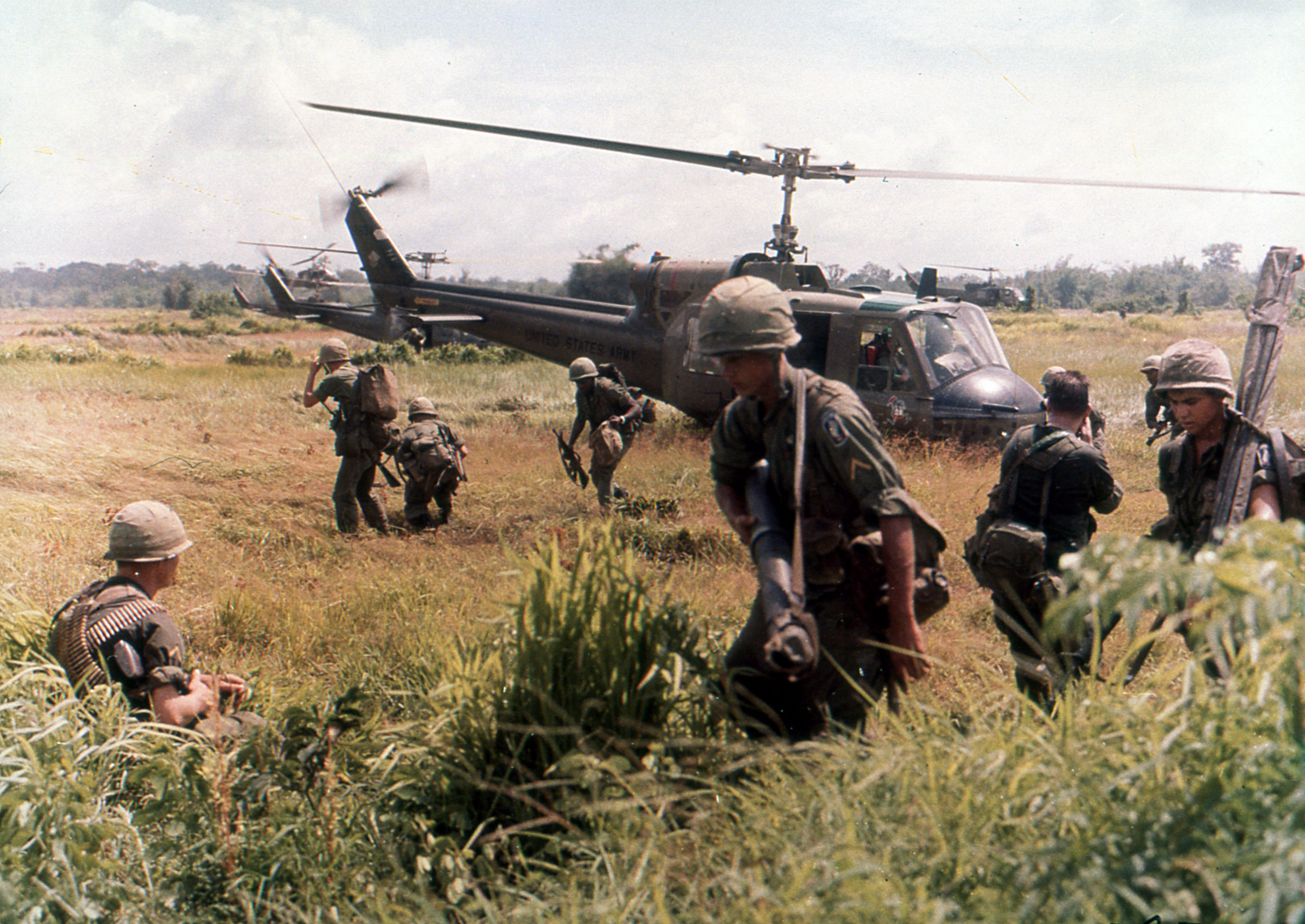 Soldiers in combat gear move through grassy terrain towards and from a hovering helicopter in a war zone