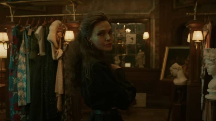 Angleina Jolie stands in a dimly lit room, wearing a dark dress, surrounded by vintage clothing and sculptures, looking over her shoulder at the camera