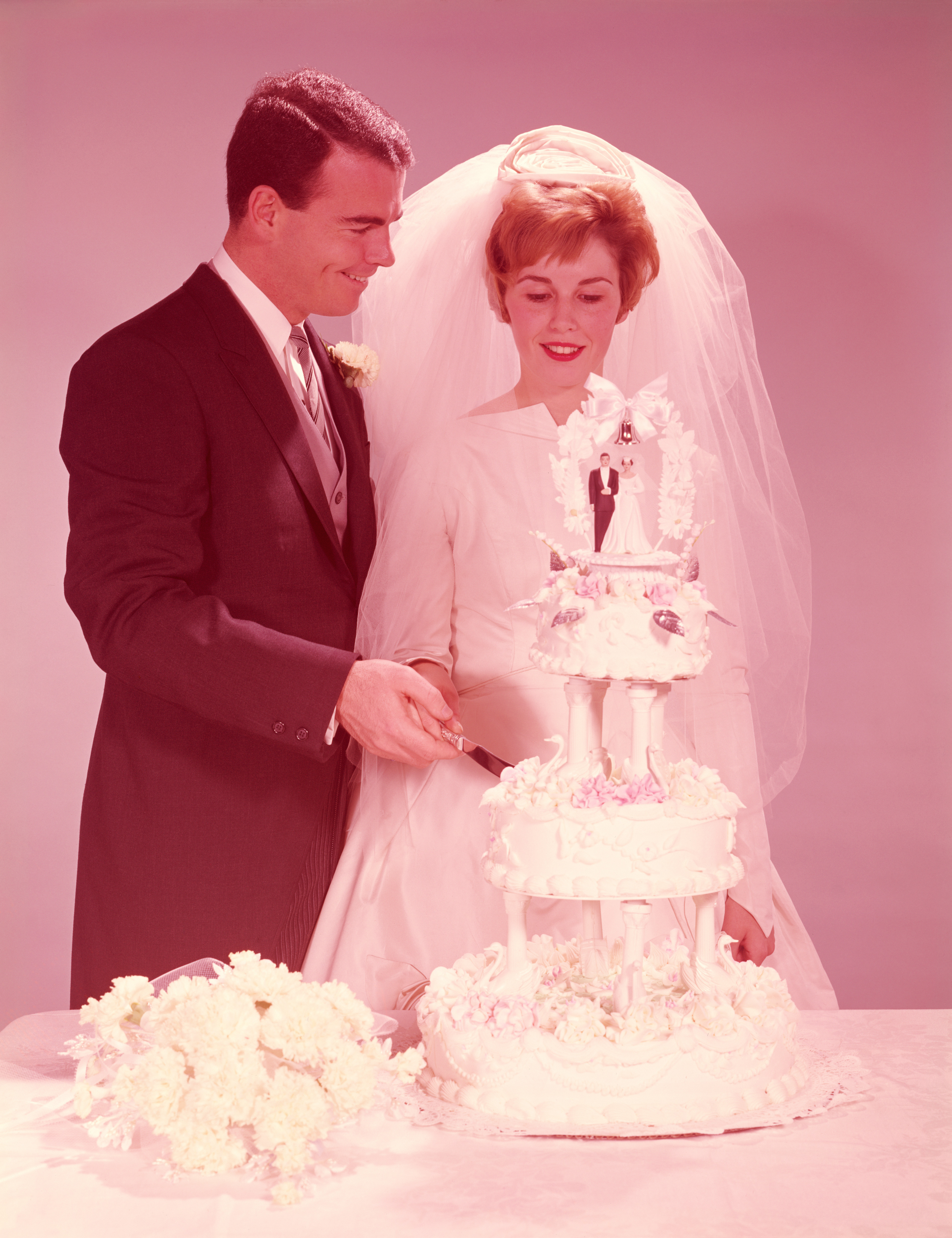 A bride in a long white dress and veil, and a groom in a black tuxedo, are cutting a multi-layer wedding cake with a bride-and-groom topper
