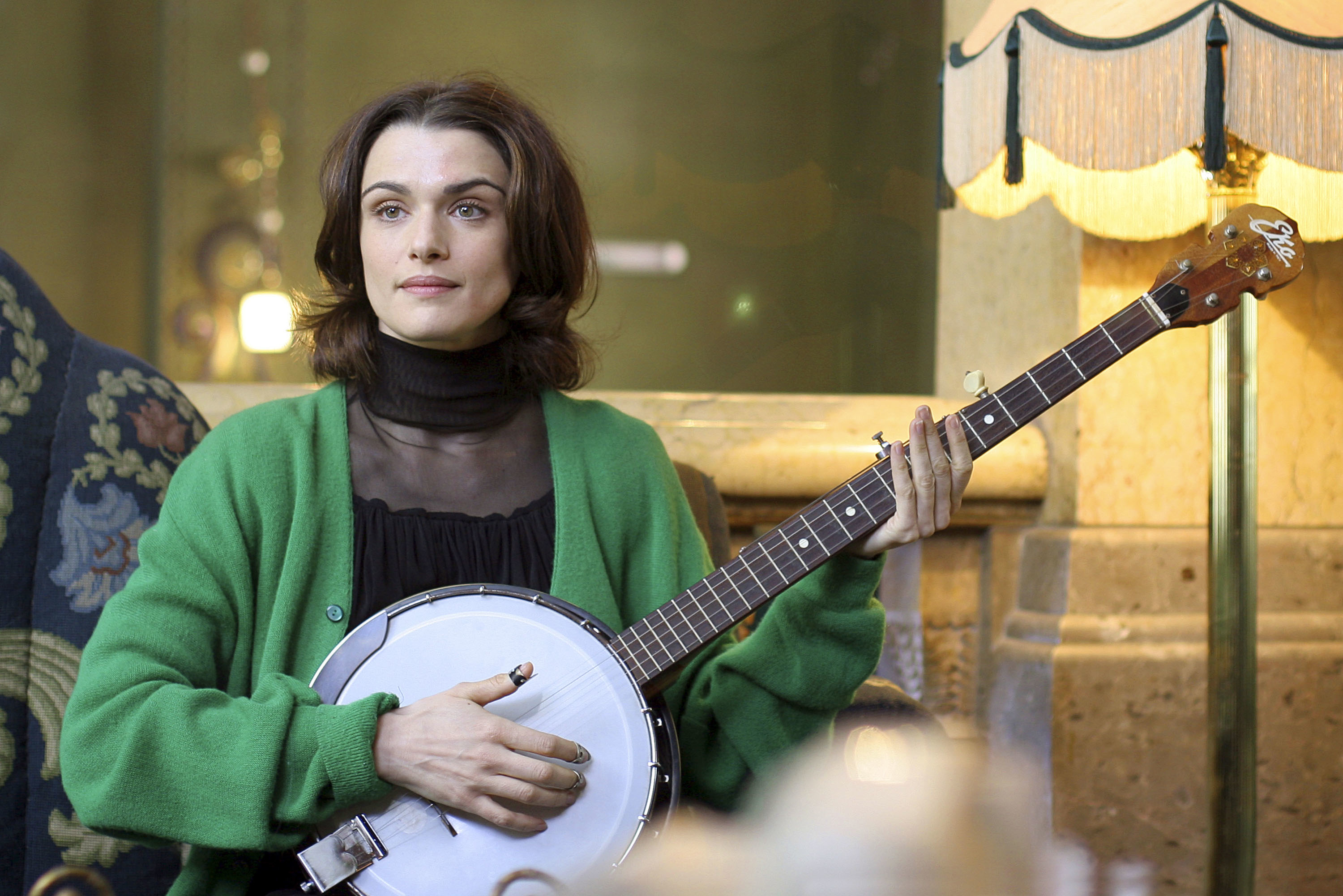 Rachel Weisz plays the banjo while seated in a room, wearing a cardigan over a dark top