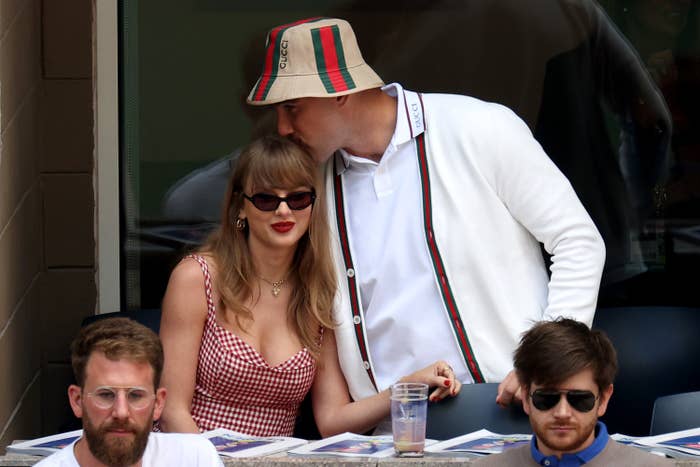 Taylor Swift in a gingham dress with sunglasses seated and listening to Matty Healy in a bucket hat, with two unidentified men in the foreground