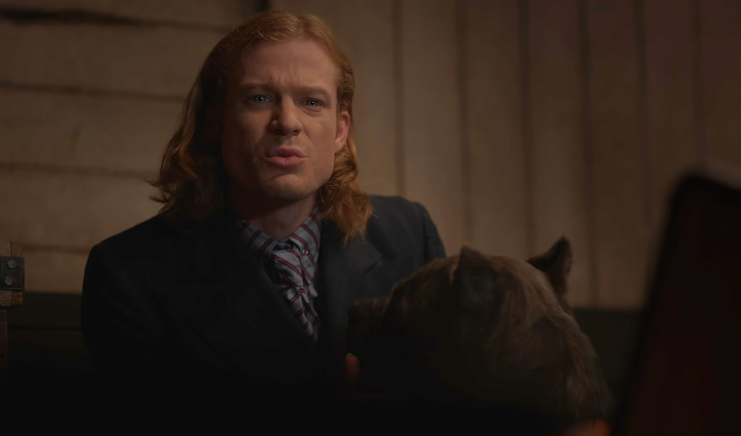 Sam Reid, wearing a formal suit, speaks seriously while next to a dog in a dimly lit room