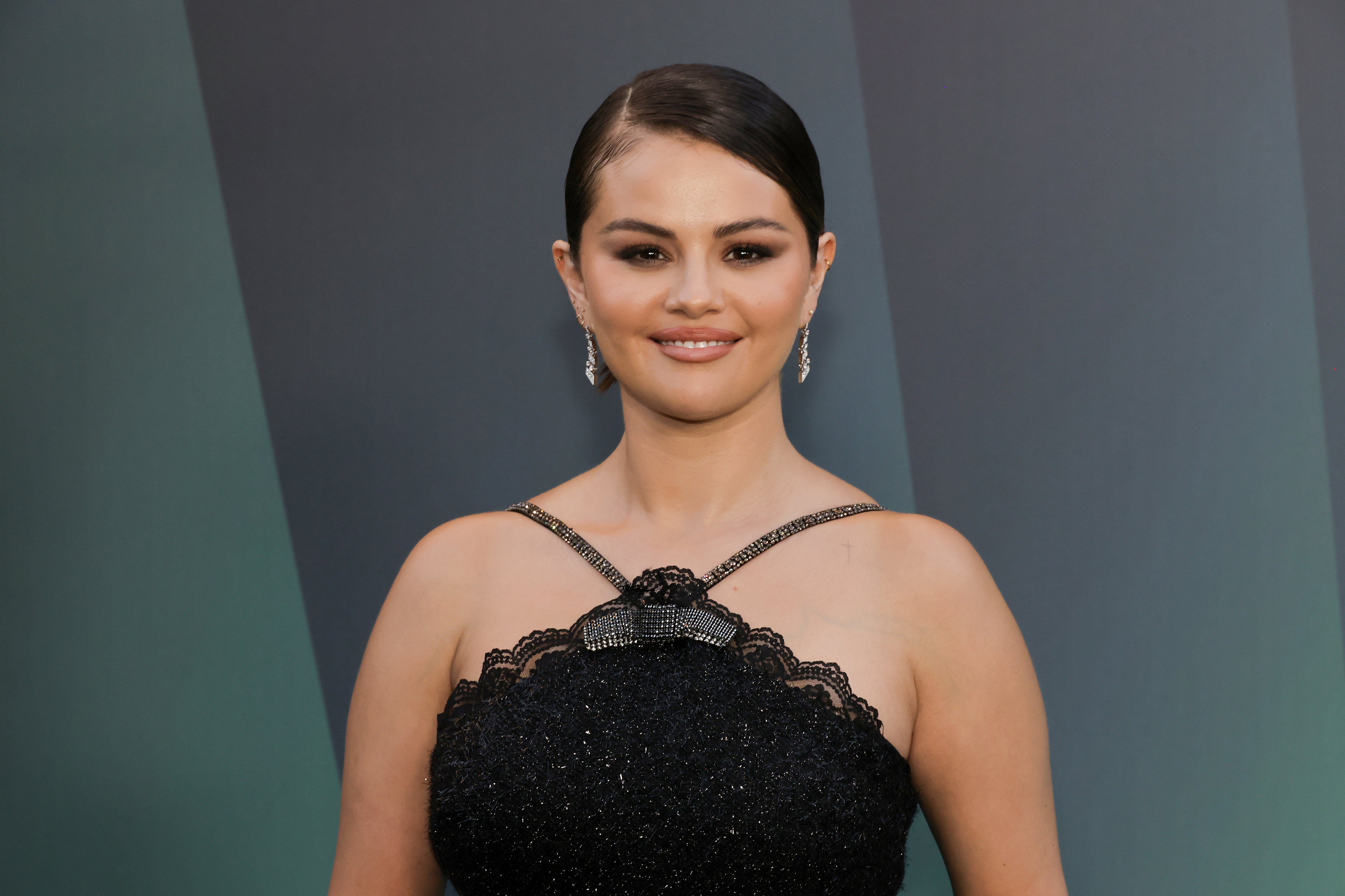 Selena Gomez, with her hair slicked back, poses in an elegant, sleeveless halter dress with beaded details and lace trim. She wears drop earrings