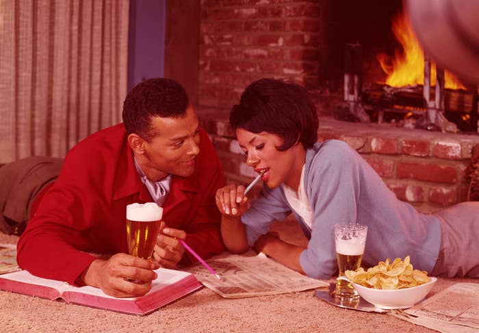 A man and a woman lie on the floor by a fireplace, reading newspapers, with drinks and snacks in front of them