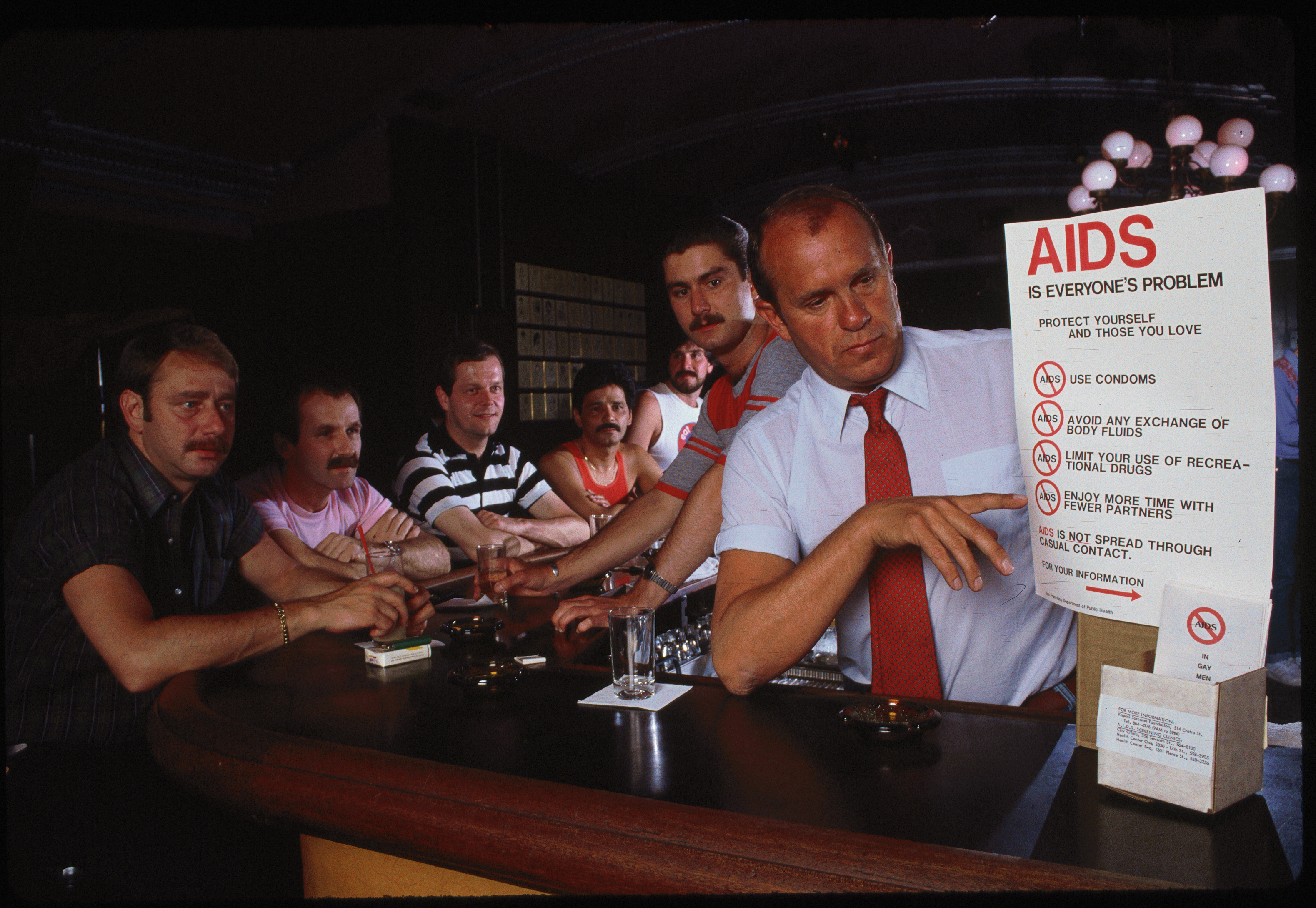Six men sit at a bar with drinks; one man in a white shirt points to an AIDS awareness sign