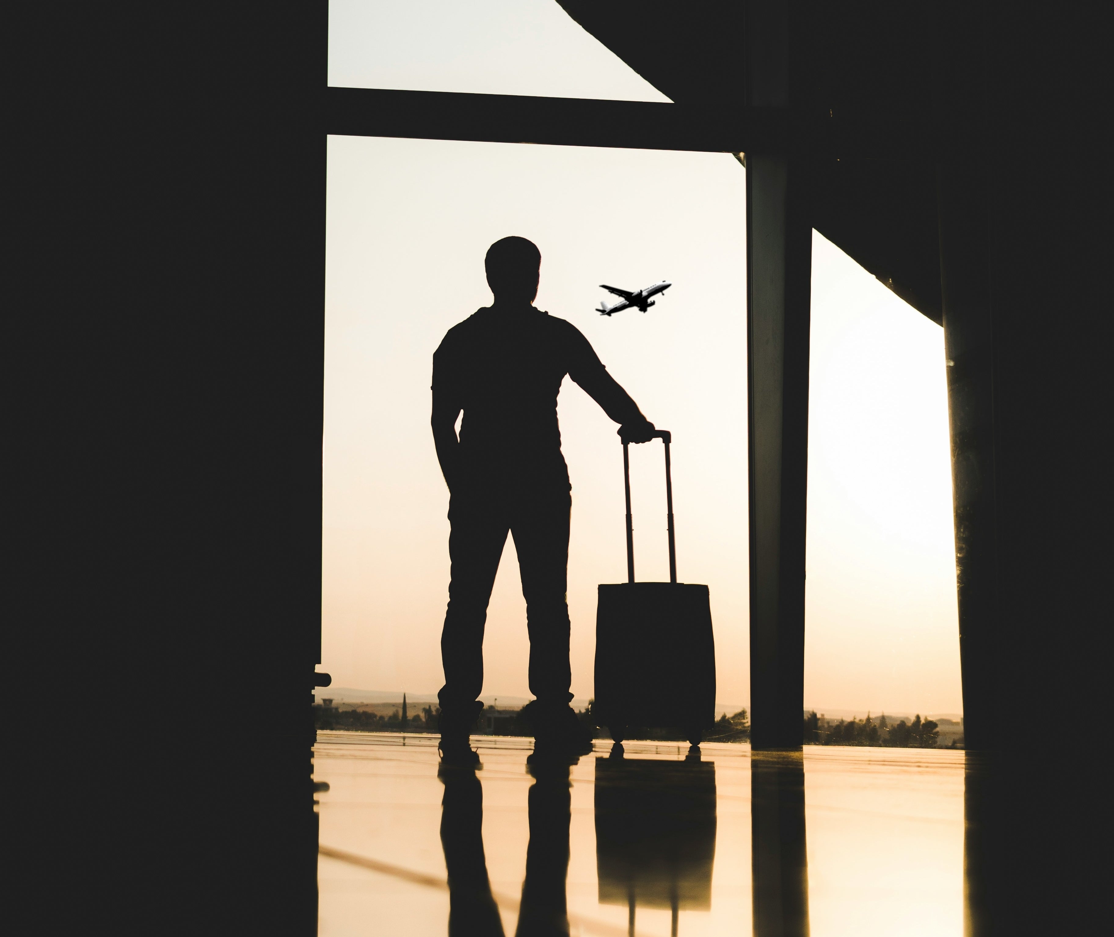 Silhouetted person standing in an airport with a rolling suitcase, watching an airplane take off through large windows