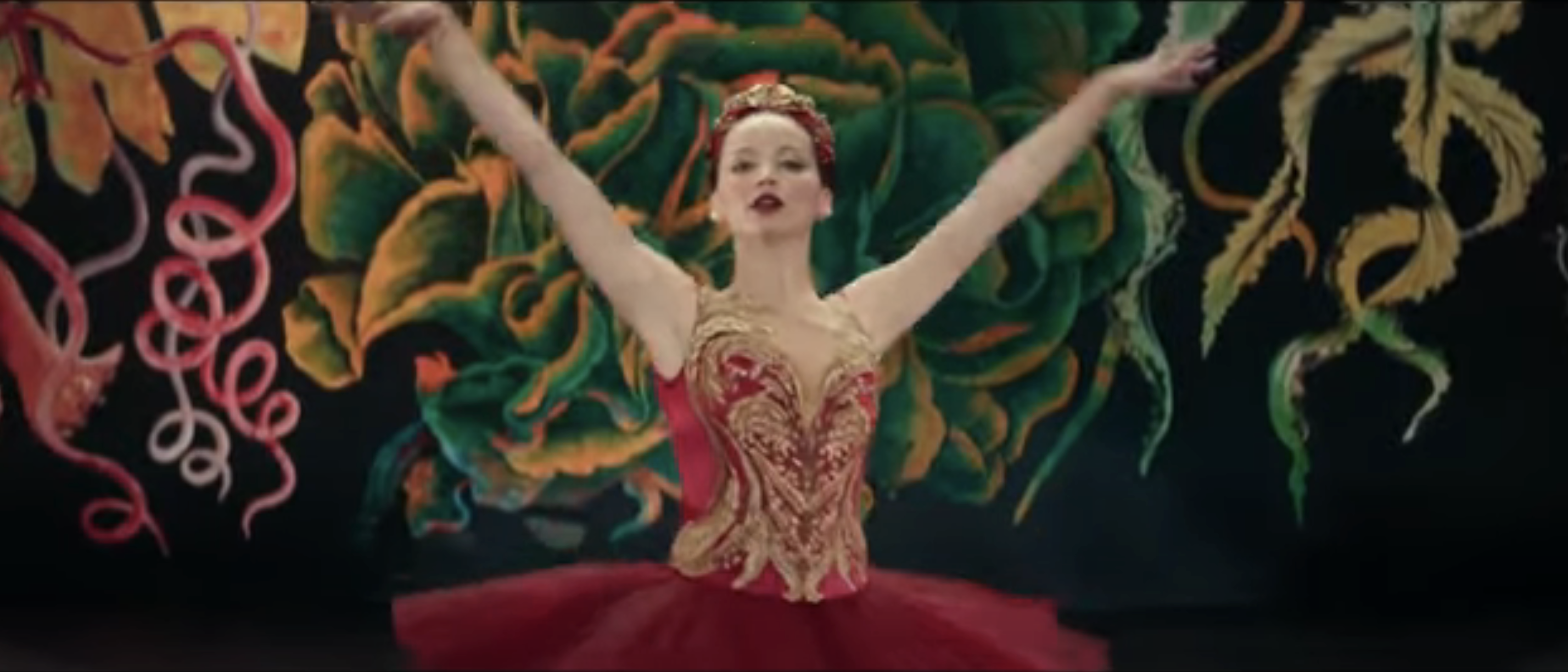 Jennifer Lawrence is dressed as a ballerina, wearing a tutu and ornate bodice, performing on stage with arms raised against a vibrant, abstract backdrop