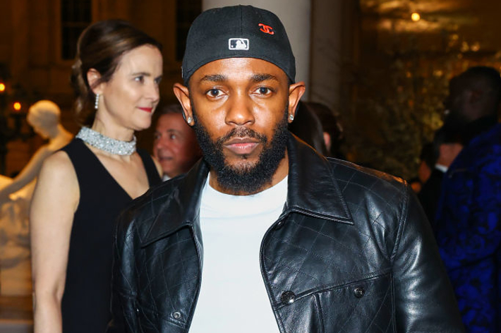 Kendrick Lamar wears a black leather jacket, a white shirt, and a red bandana as a belt, standing at an event alongside an elegantly dressed woman in a black dress