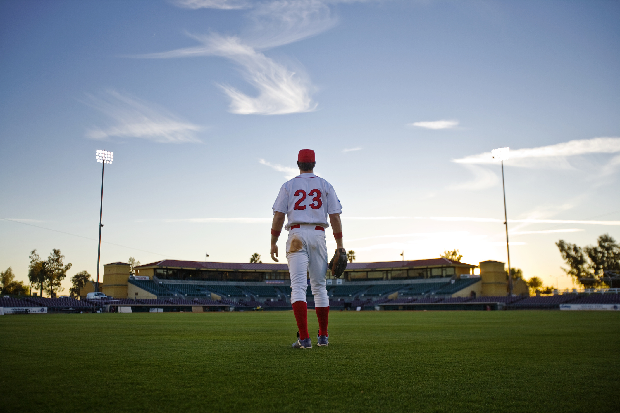 A solitary baseball player stands in a field, facing a stadium, wearing a uniform with the number 23