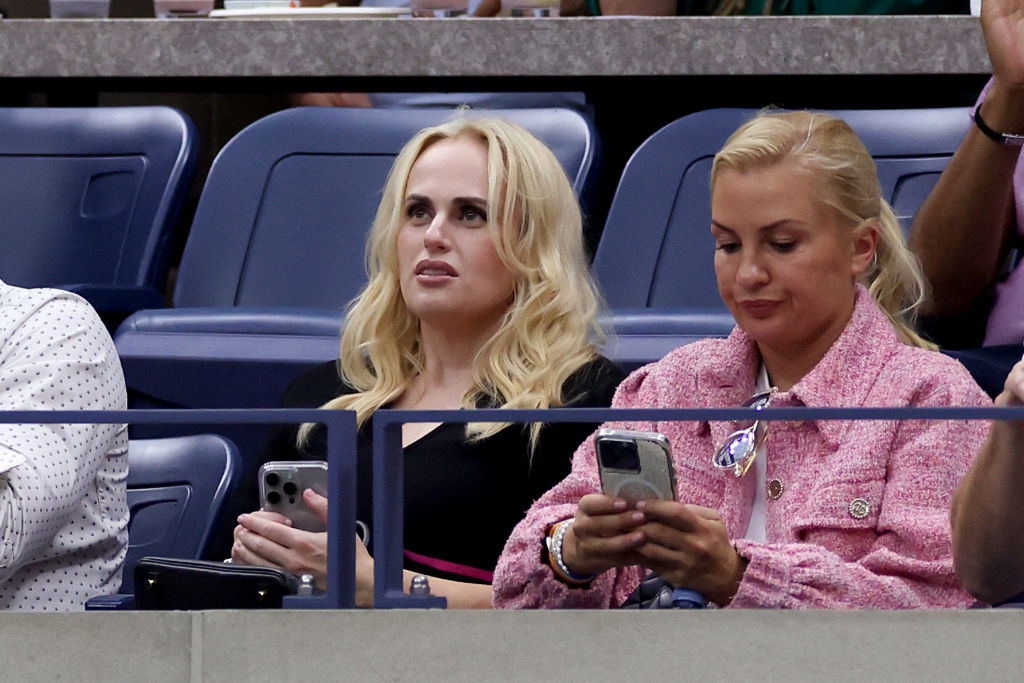 Rebel Wilson and guest sit in the stands at an event, both holding smartphones; Rebel wears a black top, and the guest wears a pink jacket