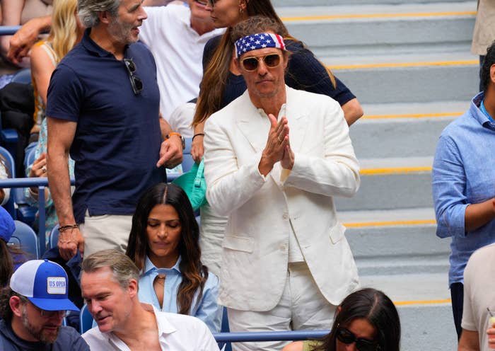 Matthew McConaughey wearing a white suit and American flag bandana, stands with hands clasped in a crowd at a sporting event. Camila Alves sits in front