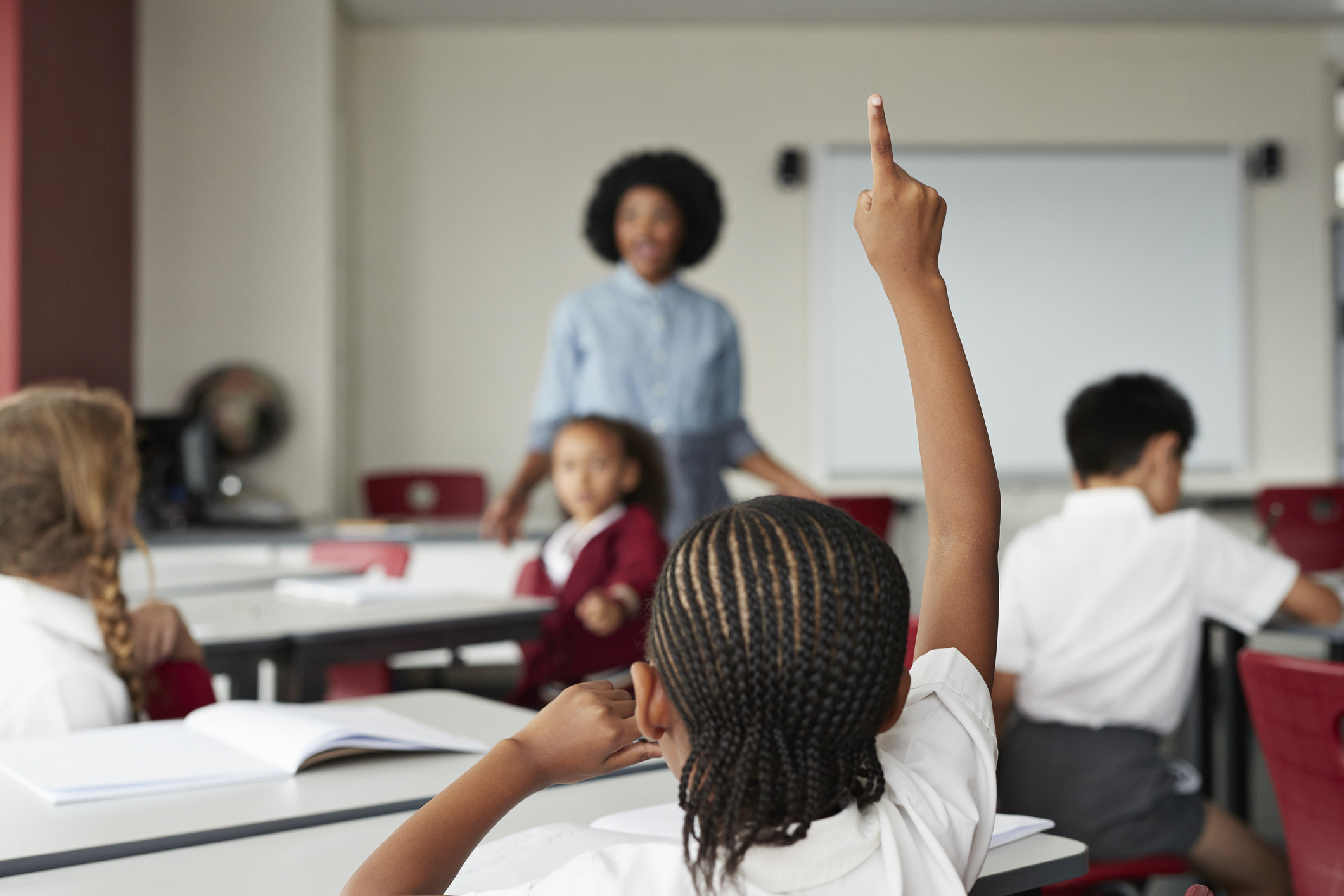 Children in a classroom, with one student raising their hand and a teacher in the background standing near the whiteboard