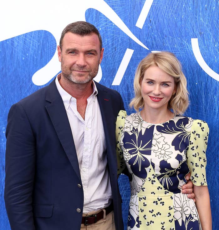 Liev Schreiber and Naomi Watts stand together at an event, both smiling. Schreiber wears a blazer and chinos, while Watts wears a floral-patterned dress