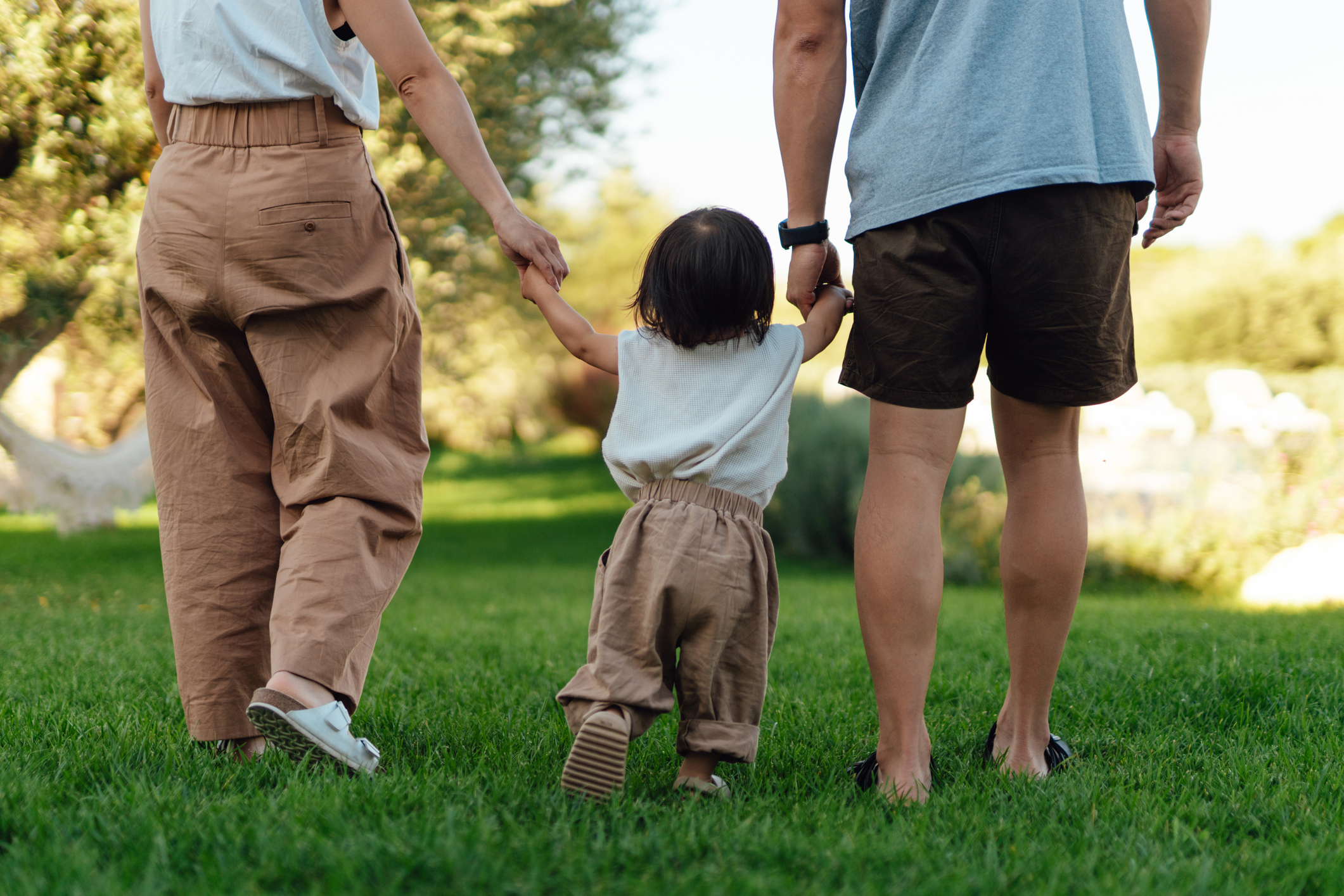 Two adults and a child holding hands and walking on grass, seen from behind