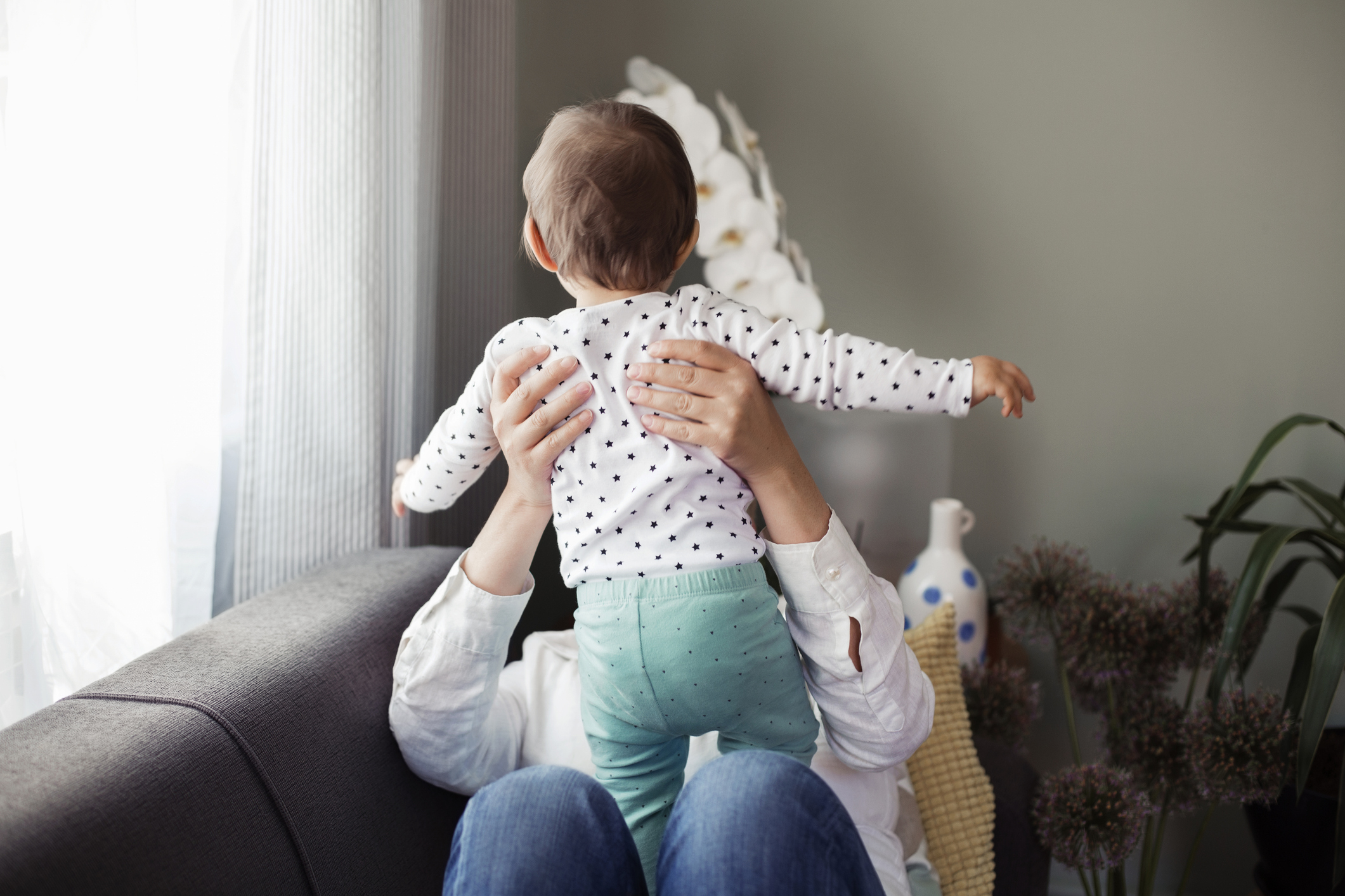An adult holds a baby with outstretched arms while seated on a sofa in a cozy living room