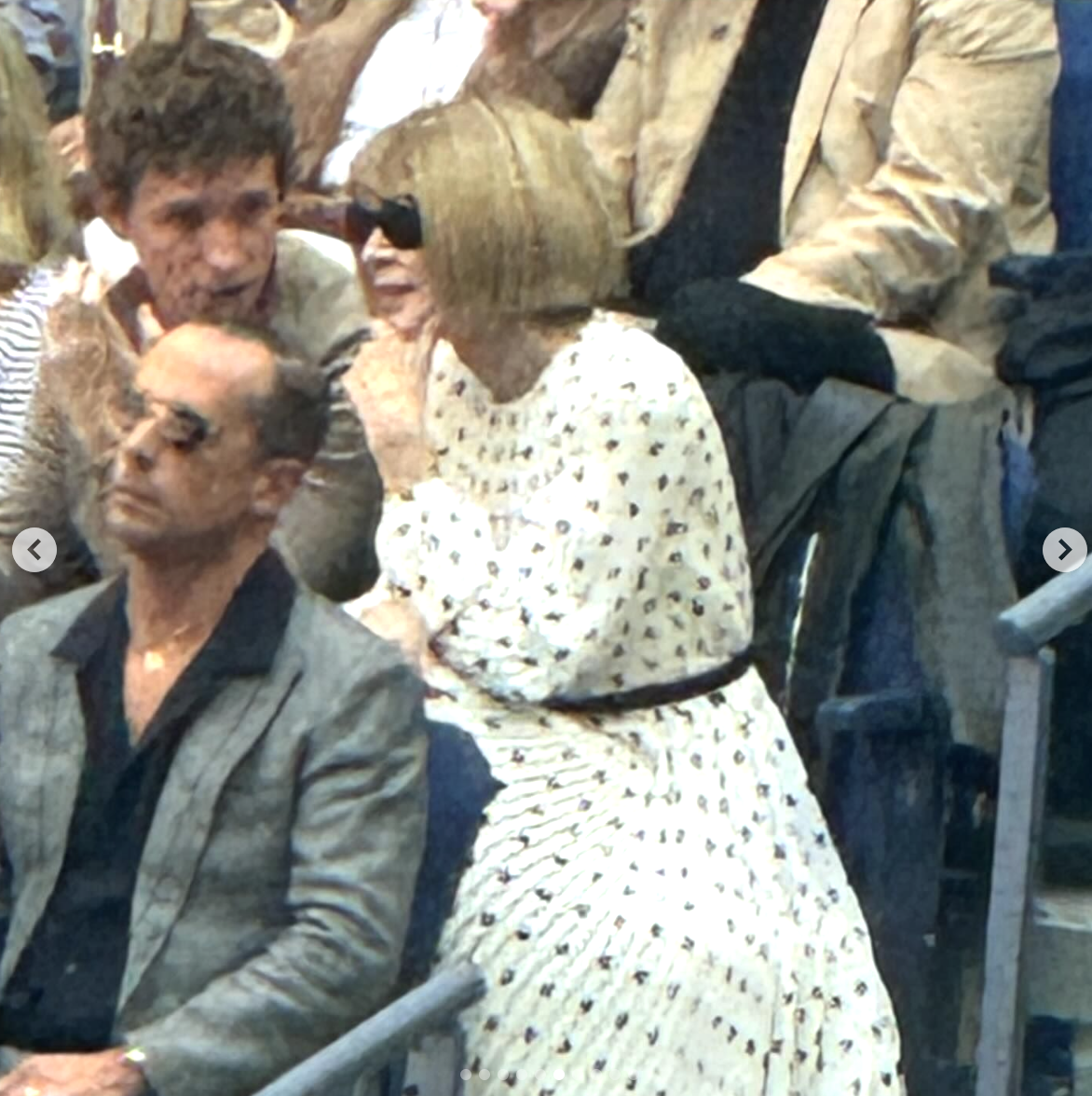 Anna Wintour in a pleated dress with a black belt speaks to a man in the audience during an event. A man in a gray blazer sits in front