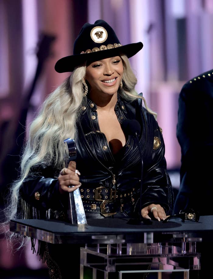 Beyoncé stands at a podium, smiling and holding a trophy, dressed in a stylish hat and leather jacket with gold details