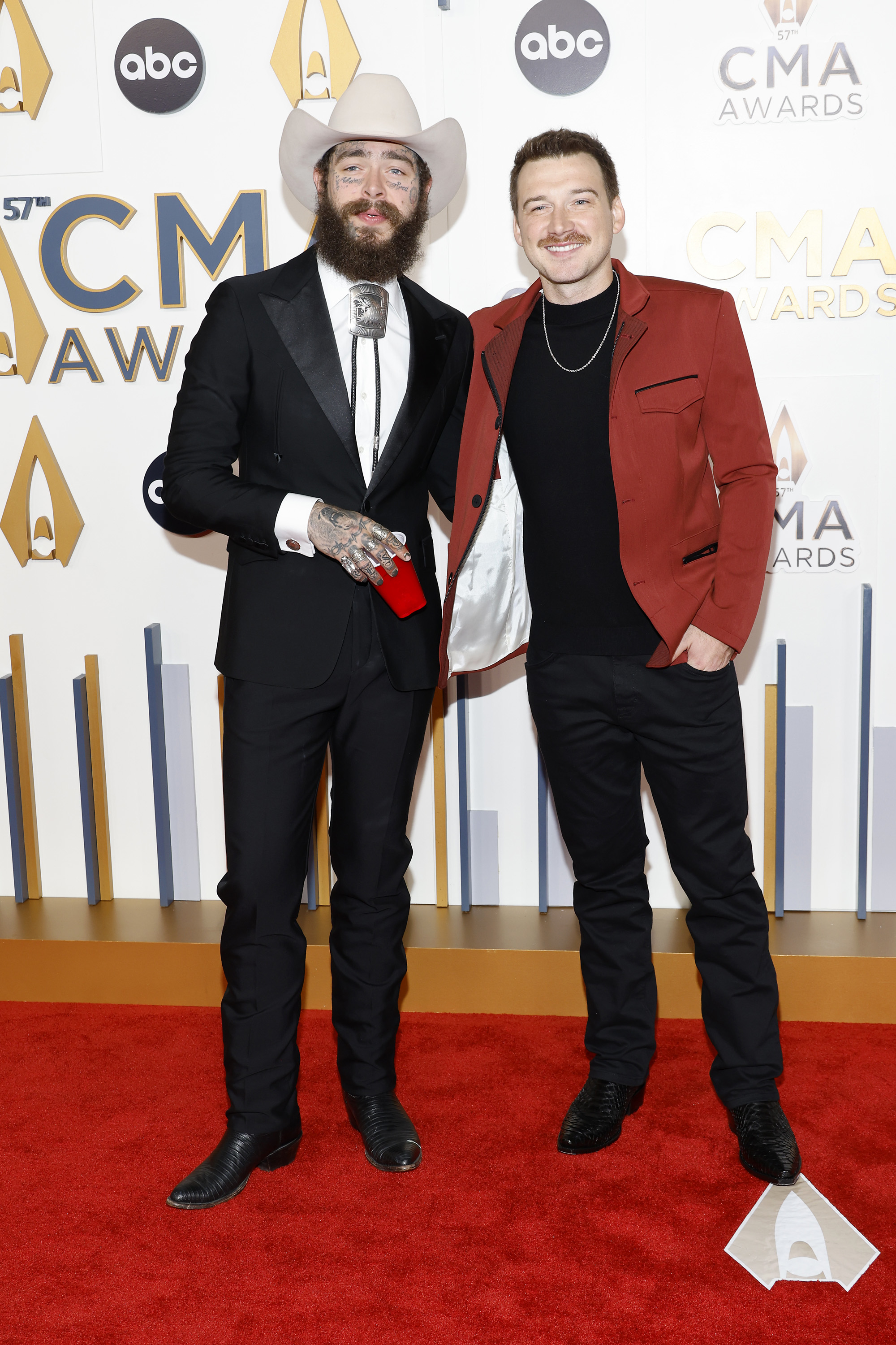 Post Malone and Morgan Wallen pose together on the CMA Awards red carpet. Post Malone is in a suit with a cowboy hat, while Morgan Wallen wears a red blazer