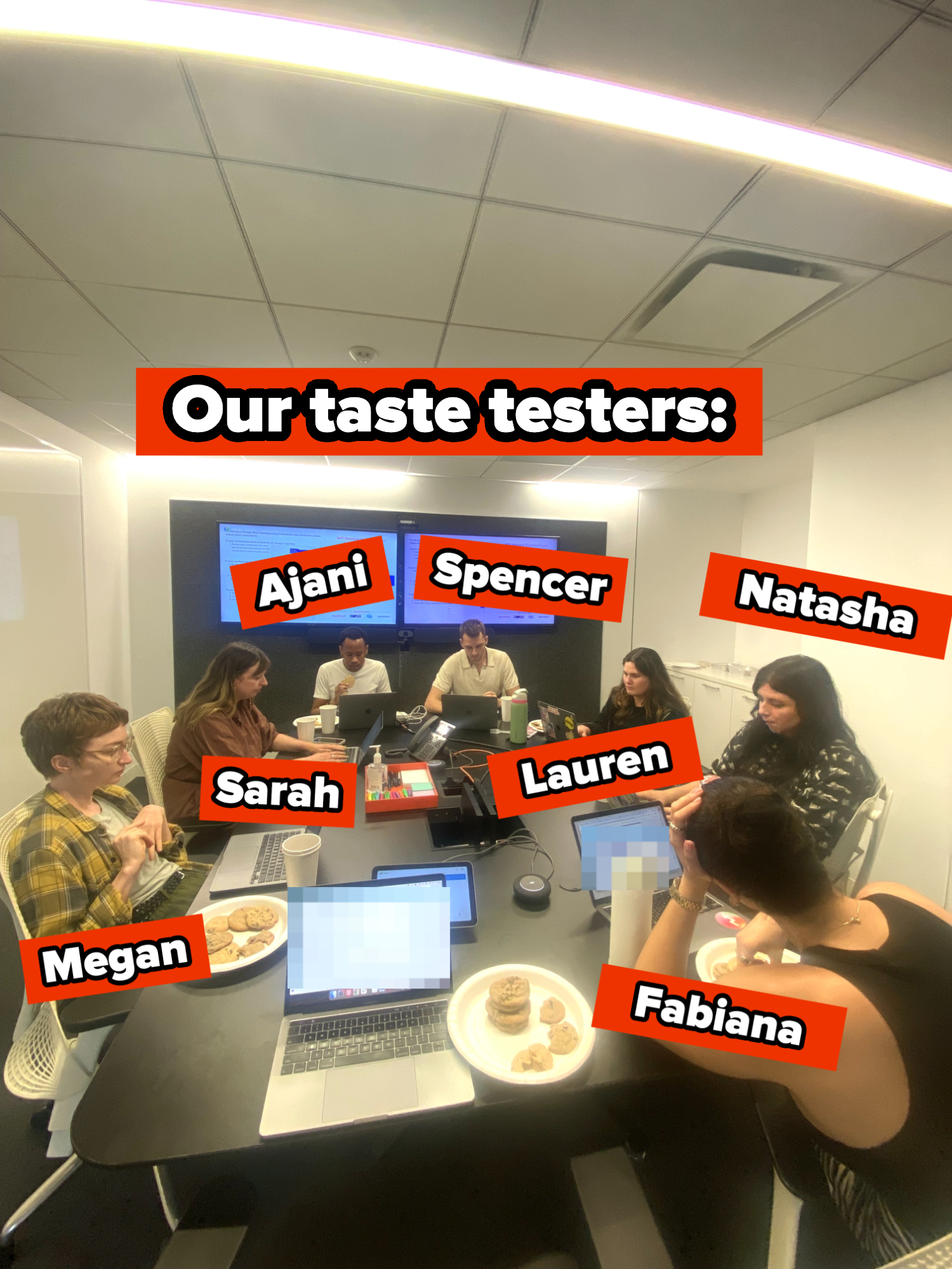 Seven people work around a conference table with laptops and plates of cookies, focusing on a collaborative project in a modern office setting