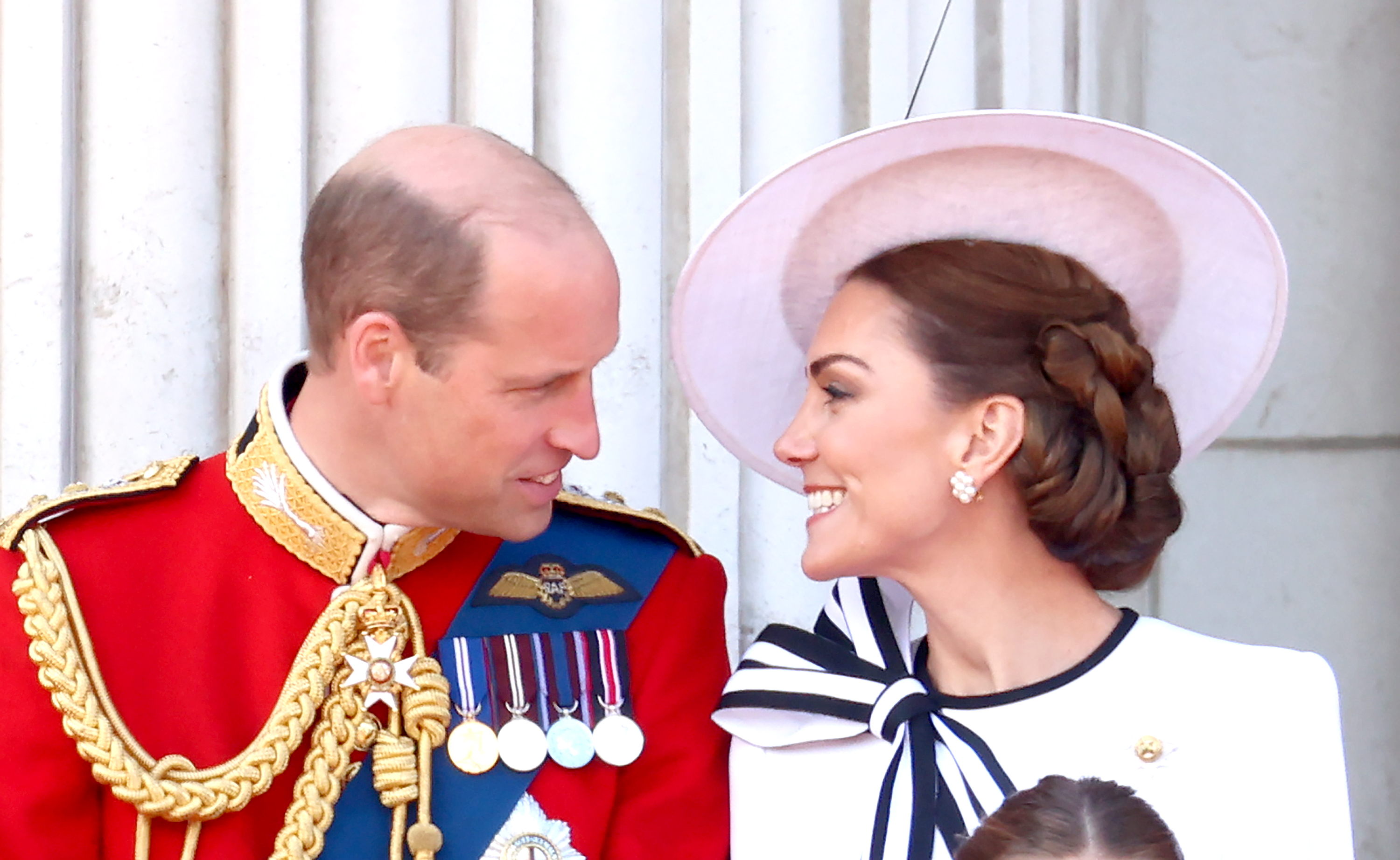 Prince William in a formal military uniform and Kate Middleton in a stylish outfit with a large hat, smiling at each other