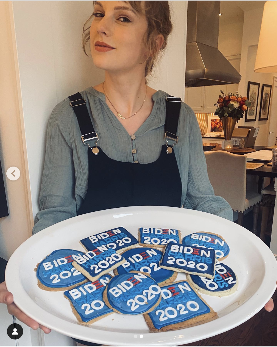 Taylor Swift holds a plate of rectangular cookies decorated with &quot;Biden Harris 2020.&quot; She is wearing a grey shirt under black overalls, posing in a kitchen