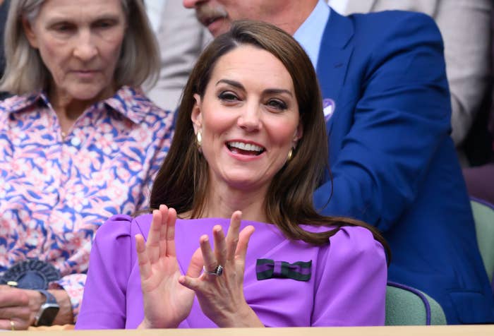 Catherine, Princess of Wales, applauds at an event. She wears a stylish dress with a bow detail on the shoulder. An older woman is seated in the background