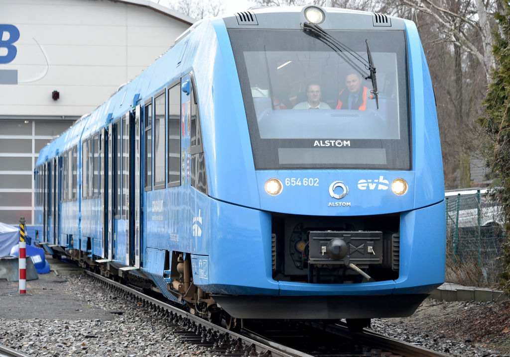 A modern blue train, labeled "Alstom," is positioned on tracks with three visible operators inside the front compartment. The train is stationary near a building